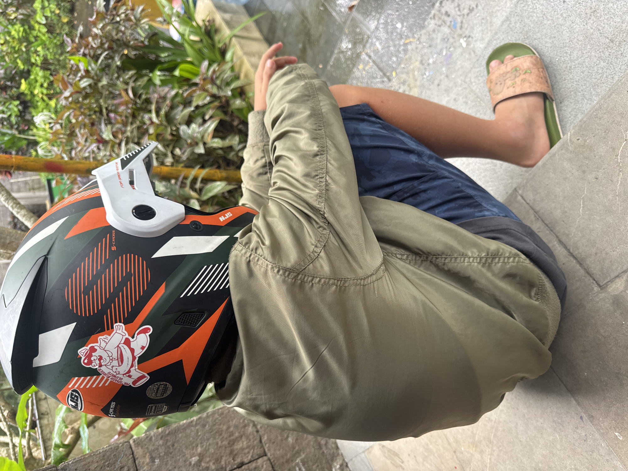A young individual wearing a military-style green bomber jacket, blue shorts, and a single green sandal is seated near some greenery and a tiled surface, seemingly outdoors. A multi-colored helmet with orange, black, white, and green designs rests nearby, decorated with text, graphics, and a cartoon sticker featuring a bear figure. The scene appears to be located in an outdoor area, possibly in Kuta Selatan, Indonesia, as suggested by the tropical greenery, although specific location markers are not visible.
