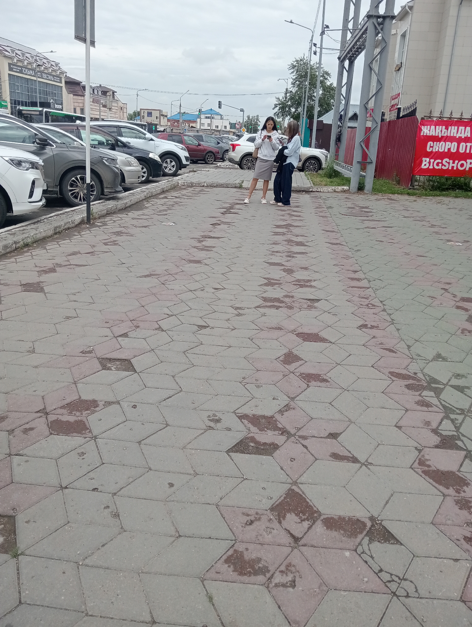 Two women stand on a sidewalk paved with hexagonal tiles. The woman on the left wears a white hoodie and a grey skirt, holding a smartphone. The woman on the right wears a light blue jacket and dark pants, also looking at a smartphone. Behind them, a row of parked cars is visible, including white, grey, and dark-colored vehicles. In the background, buildings and streetlights indicate an urban setting. A red banner with white text is affixed to a fence on the right side of the image. The sky is overcast. This scene is located in Kostanay, Kazakhstan, identifiable by the sign reading "ЖАҚЫНДА СКОРО ОТКРЫТИЕ BIGSHOP" on the red banner, which is in Kazakh and Russian, and a sign on a building in the background reads "ЮКОМА ТЕКСТИЛЬ," also common in Kazakhstan.