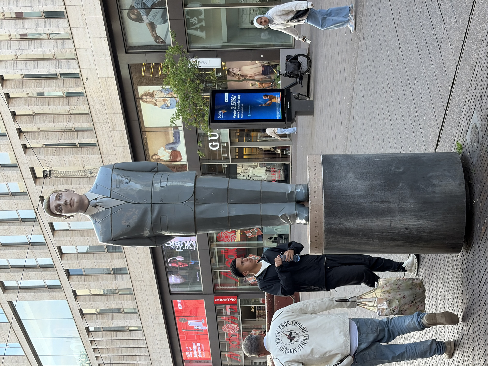 This image depicts a daytime street scene on Spui, a pedestrian shopping street in The Hague, Netherlands.
In the central foreground, a large, segmented statue of a male figure stands atop a dark, cylindrical pedestal. The statue, identified as "De Waarnemer" (The Observer), is light grey/blue, depicting a man in a suit and tie, facing forward. Horizontal lines segment the statue's body. The pedestal's base is inscribed with "DE WAARNEMER" and "THE OBSERVED" (partially visible). The ground is paved with rectangular grey and brown interlocking bricks.
Three individuals are visible in the mid-ground. To the left, a man with grey hair, seen from behind, wears a light-colored jacket with a black skull and text graphic on the back ("KRSH ENGRO BRAND SINCE 01 UNLIMITED"). He wears light blue jeans and light-colored shoes, carrying a floral-patterned shopping bag in his left hand. To his right, another man, facing slightly right, wears a black hooded jacket, white t-shirt, black trousers, and white athletic shoes, holding a clear plastic water bottle. Further right, a woman in a white headscarf (hijab), light-colored sweater, light blue wide-leg jeans, and light athletic shoes walks towards the left, carrying a black shoulder bag and holding a small white object in her right hand. Near the entrance of the "GUESS" store, a second woman, seen from behind, stands in light clothing.
The background features multi-story commercial buildings. The ground floor displays several retail establishments: "SONY" with a promotion featuring a man in headphones and "For The Music," a partially visible "HEAT-WAVE" sign, a "Metro&Mani" sign, "GUESS" with mannequins in the window, and "A&F" (Abercrombie & Fitch). A vertical digital billboard near the "GUESS" store displays a Dutch advertisement for "Bunq" bank, offering "2,51% rente" for "6 maanden lang," featuring a person holding a blue stick. A black bicycle is parked behind the billboard, alongside a small green tree. The upper floors of the main building feature a light-colored, panel-clad facade with numerous tall, narrow rectangular windows reflecting the sky and surrounding structures. Overhead wires, indicative of tram lines, traverse the upper portion of the scene.