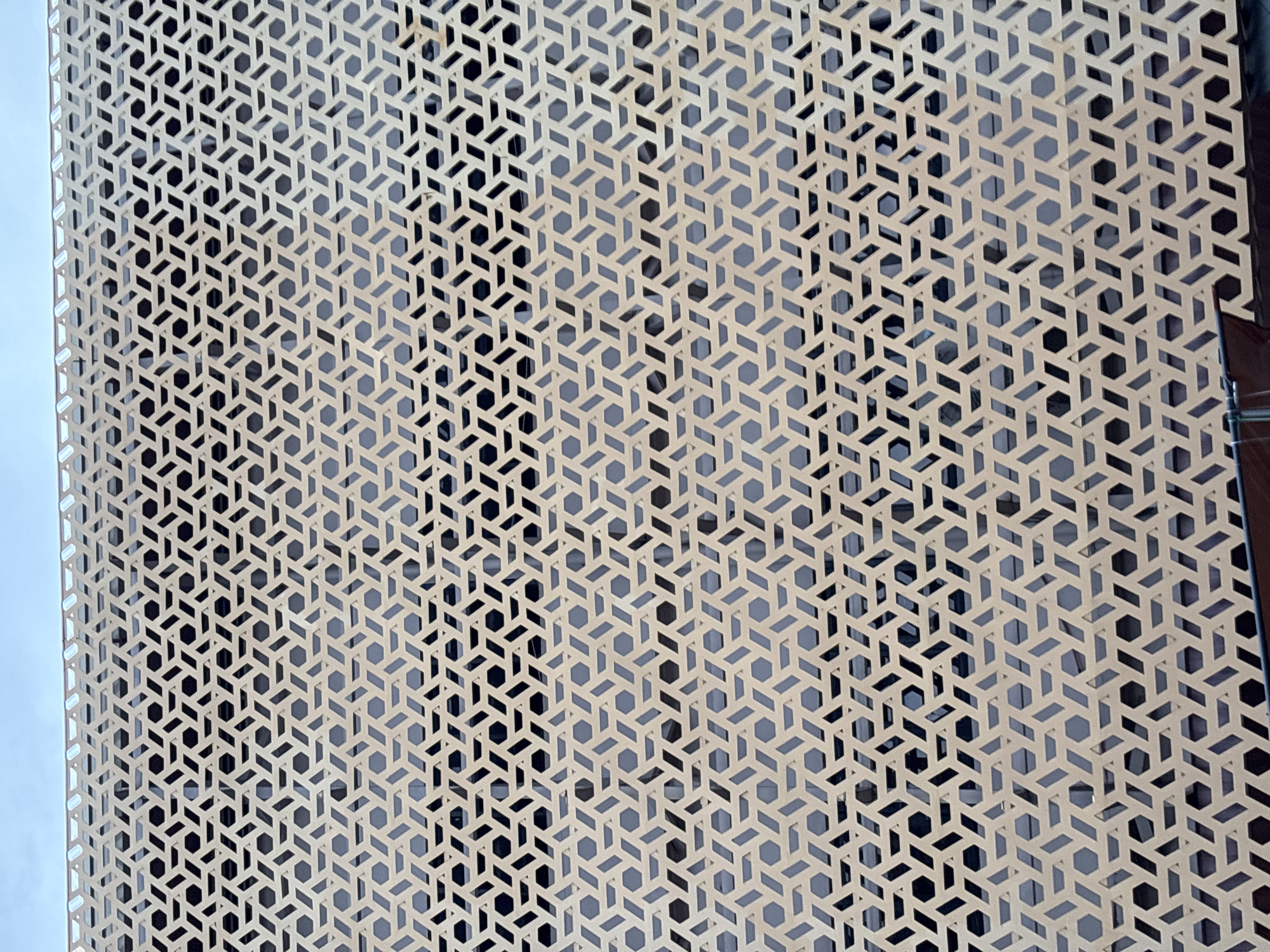 The media file shows a close-up view of a decorative screen or facade with an intricate geometric pattern. The pattern is composed of repeating hexagonal and rhomboid shapes, creating a lattice-like effect. The screen is a light beige or tan color and appears to be made of metal or a similar durable material. Behind the screen, darker shapes are visible through the cutouts, suggesting it is mounted on a building or structure with windows or a darker backing. The lighting suggests it is daytime, possibly outdoors, given the bright exposure and potential for ambient light. The scene is devoid of people or any overt activity, focusing solely on the architectural detail. There is no visible text. The location context is Davao City, Philippines, which might imply a tropical climate, but this cannot be definitively determined from the image alone. The repeating pattern is the most prominent feature, highlighting a modern or possibly traditional-inspired design element.