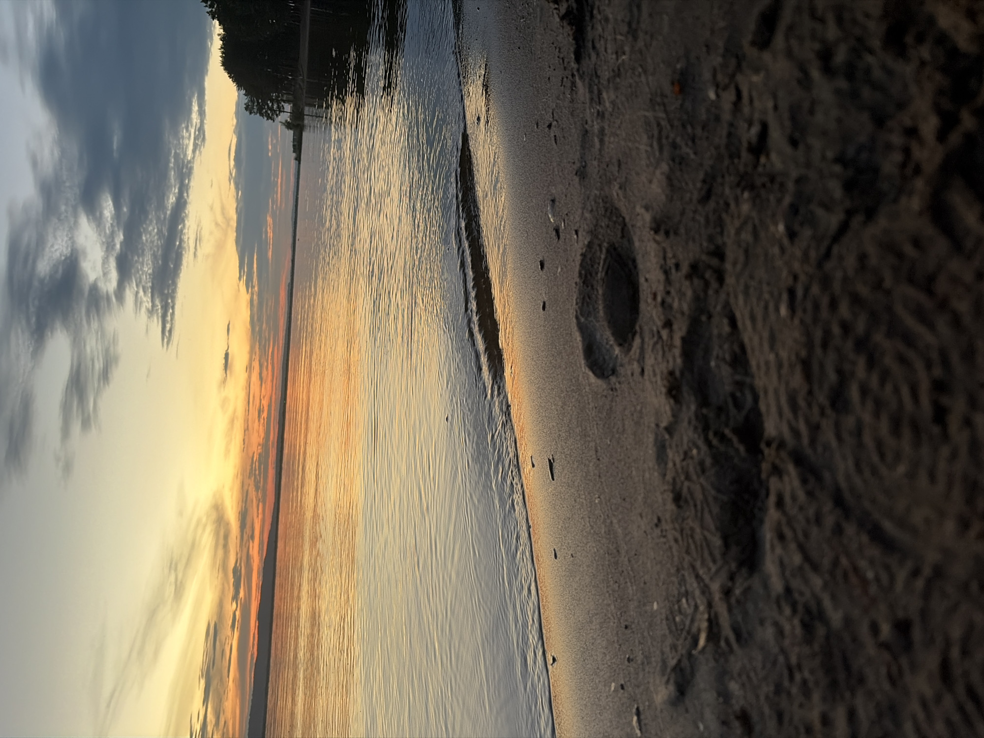 This media file depicts a serene natural scene at the edge of a body of water, likely a lake or ocean, during a sunset. The image is oriented vertically, with the foreground dominated by sand and the background by water and sky.
The sand in the foreground is textured, with visible footprints and imprints, suggesting recent human presence. It transitions to the water's edge, where small waves are gently lapping the shore. The water itself reflects the warm hues of the sunset, creating a shimmering effect with ripples and lines across its surface.
The sky displays a gradient of colors characteristic of a sunset. Dark, dramatic clouds occupy the upper left portion, gradually giving way to lighter shades of yellow and orange closer to the horizon. The horizon line is marked by a distant shoreline with trees, silhouetted against the vibrant sky.
The time of day is clearly sunset, indicated by the warm, low-angled light and the colors in the sky and water. The overall mood conveyed is peaceful and tranquil, evoking a sense of calm. The location appears to be a natural beach or shoreline. There is no visible text in the image.