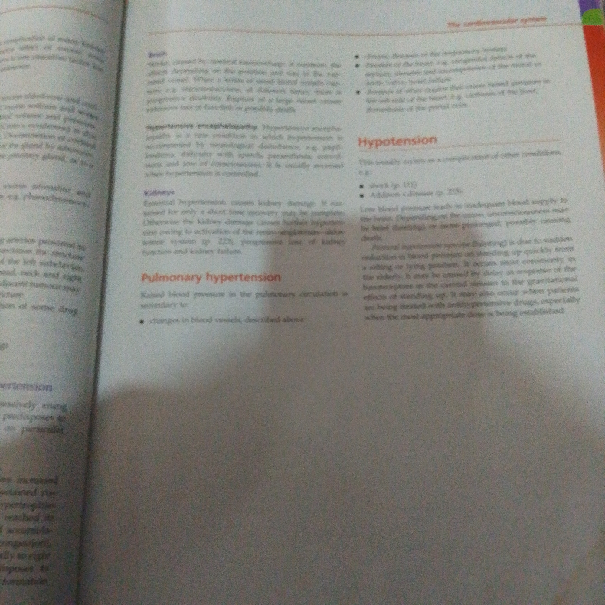 The image displays an open book with text about medical conditions related to the cardiovascular system. The book is lying flat, suggesting it is being read or referenced. The lighting appears to be artificial, possibly from an overhead lamp or room lighting, as there are no natural light cues. The text is predominantly in English and includes headings such as "Hypotension," "Pulmonary hypertension," and "Kidneys." The subjects are the printed words and layout of the book itself, rather than people or a specific scene. The activity is informational, focused on the presentation of medical knowledge. There are no discernible location cues within the image that specifically point to Jalingo, Nigeria, or any particular time of day or weather. The overall impression is one of academic study or reference material.