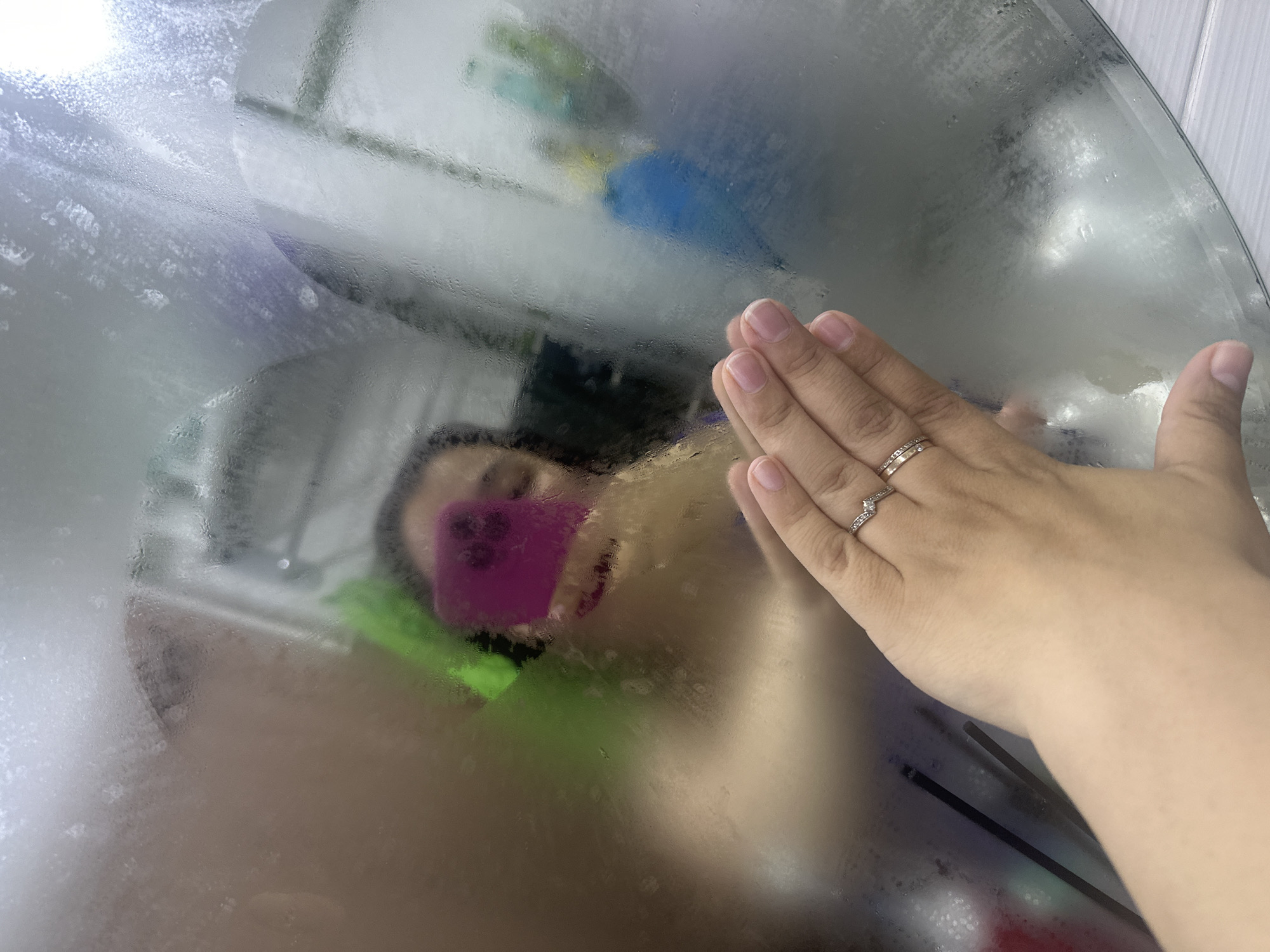 A close-up, first-person perspective image depicts a human hand touching a circular or oval mirror covered in condensation. The hand, positioned in the lower-left foreground, displays two rings on the ring finger: an upper silver-toned band with a V-shaped pattern of small stones, and a lower gold-toned band, also possibly with small embedded stones. Fingernails are neatly manicured.
Through the translucent fog and the clearer area where the hand makes contact, a blurred reflection of a person is visible. This reflection shows an individual, likely an adult female, with dark hair, holding a bright magenta/purple smartphone up to capture the mirror reflection. The person's face is largely obscured by the phone and the fog. To the immediate left of the reflected person's head, a blurred bright green object is discernable. To the right, a blue object, possibly a container, is visible. Other indistinct shapes and light sources are present in the background reflection, consistent with a bathroom setting. The mirror itself shows various patterns of condensation and water droplets. Below the mirror's edge in the bottom-right, a light-colored, possibly tiled or paneled, wall is visible.
Location: Unknown, Unknown.