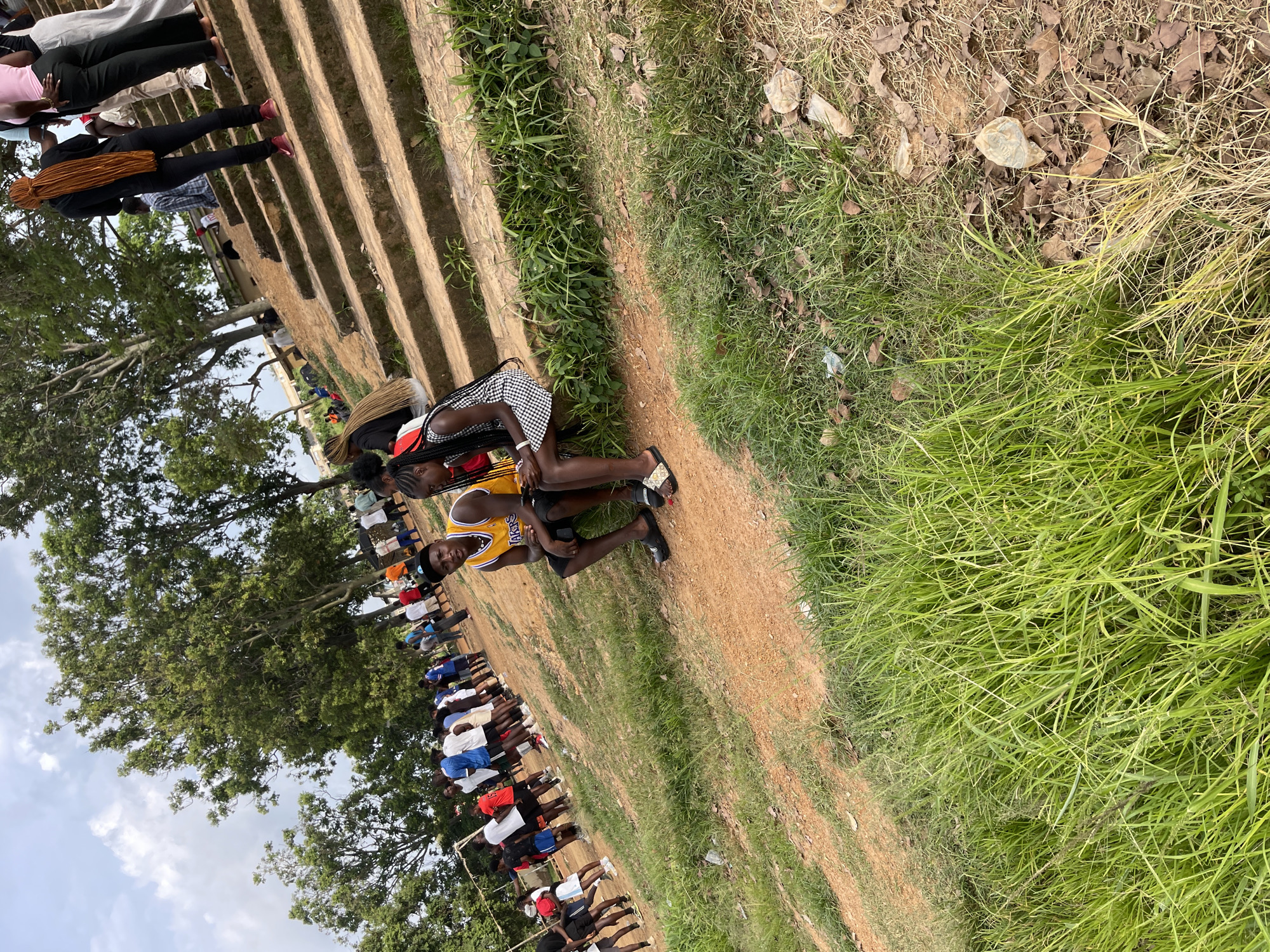 In Rafin Par, Nigeria, on an outdoor, uneven dirt and grass terrain with a set of concrete stairs in the background, two young adults, a male and a female, are seated on the edge of the stairs. The male, on the left, is wearing a yellow basketball jersey with the name "Lakers" and black shorts. The female, on the right, is wearing a black and white checkered dress. A crowd of people, predominantly young adults, are visible in the background, some standing on the stairs and others on the dirt area. The environment is characterized by lush green grass in the foreground and scattered dry leaves on the ground. Trees with dense foliage are present in the upper background, and the sky is partly cloudy. The scene appears to be a casual gathering or event.