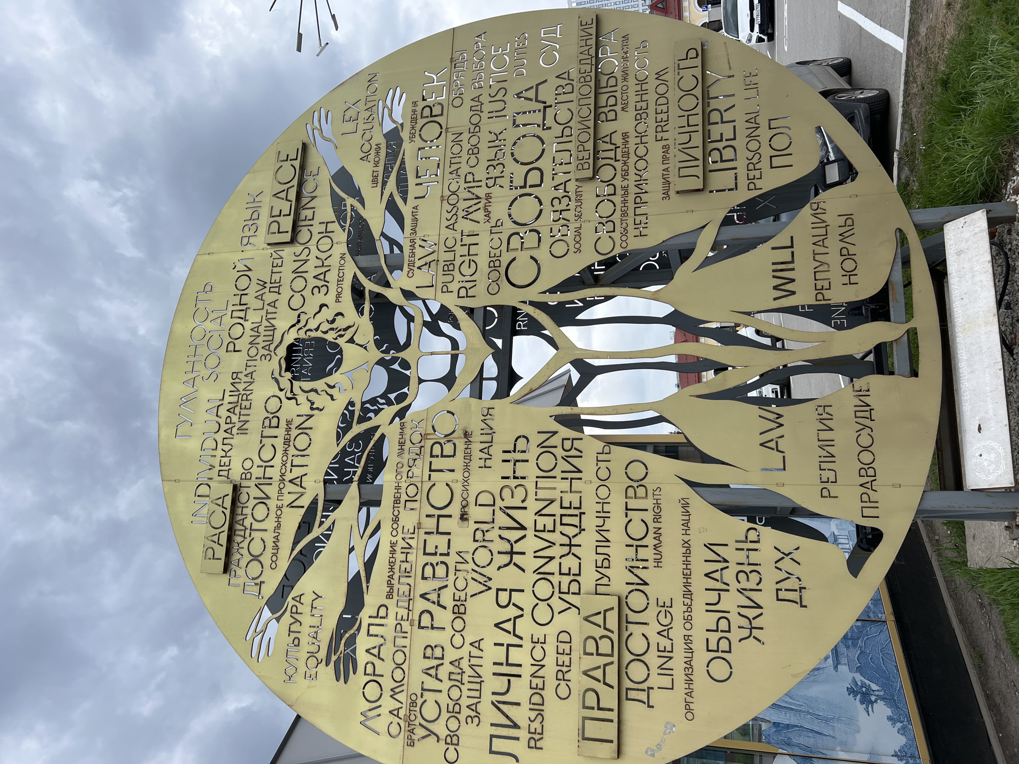 A low-angle shot showcases a circular, golden metal monument in Ufa, Russia. The monument is engraved with a stylized human figure formed from the words of the Universal Declaration of Human Rights in Russian and English. The words are arranged to create the form of a tree and a human figure. The monument sits on a gray metal stand with a white rectangular sign attached to the side. The background includes a cloudy sky, a building, cars, and some greenery.