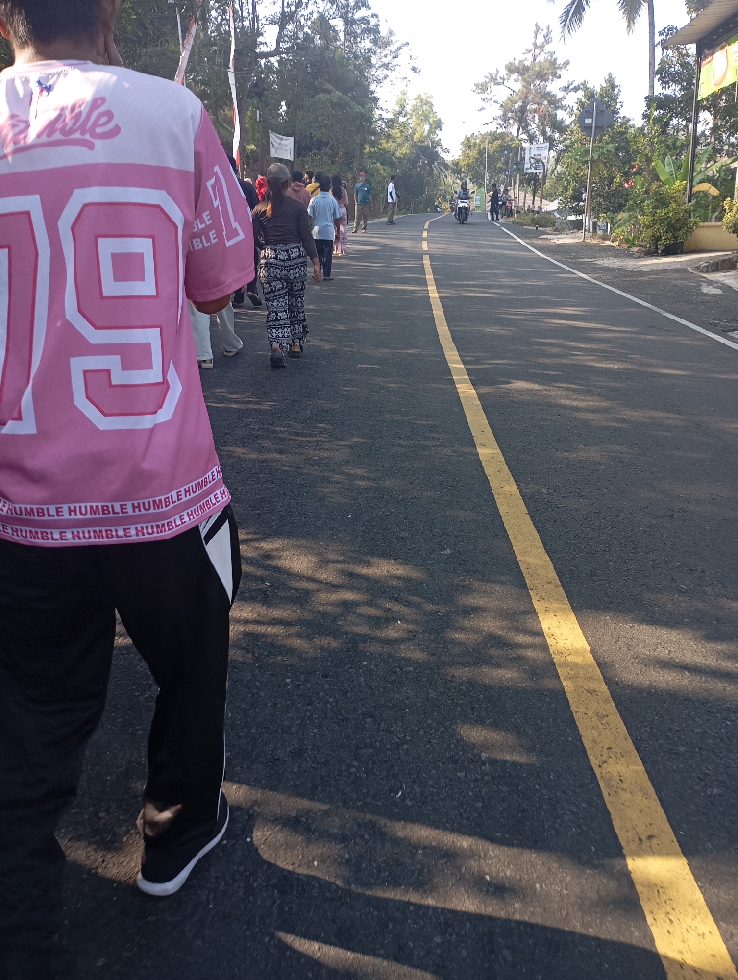 A group of people are walking along a paved road in Ketepung, Indonesia. The road has a yellow line down the center and is bordered by trees and foliage. In the foreground, a person wearing a pink jersey with the number 19 on it and black pants is walking away from the camera. Other individuals are visible further down the road, some appearing to be walking in groups. There are also a few motorcycles present on the road. The lighting suggests it is daytime, with sunlight casting shadows on the asphalt.