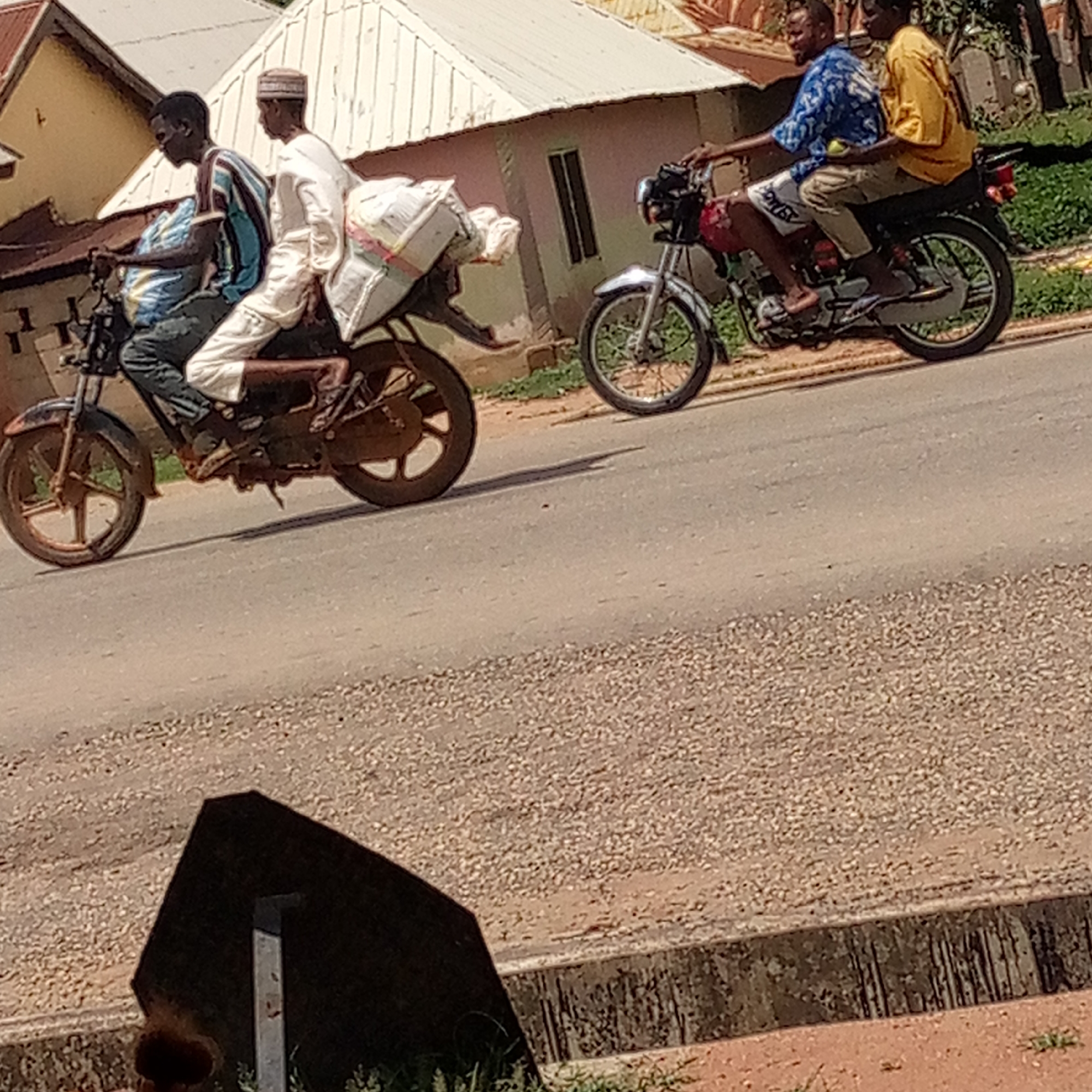 The image captures a street scene in Mararaba, Nigeria. Two motorcycles are traveling on an asphalt road. The first motorcycle carries three people, the driver, a passenger in the middle, and a third person sitting on the back of the seat holding a bundle. The second motorcycle has two passengers, the driver and a person seated behind. The background includes buildings with corrugated metal roofs. In the foreground, there is a metallic object or structure and a concrete curb.