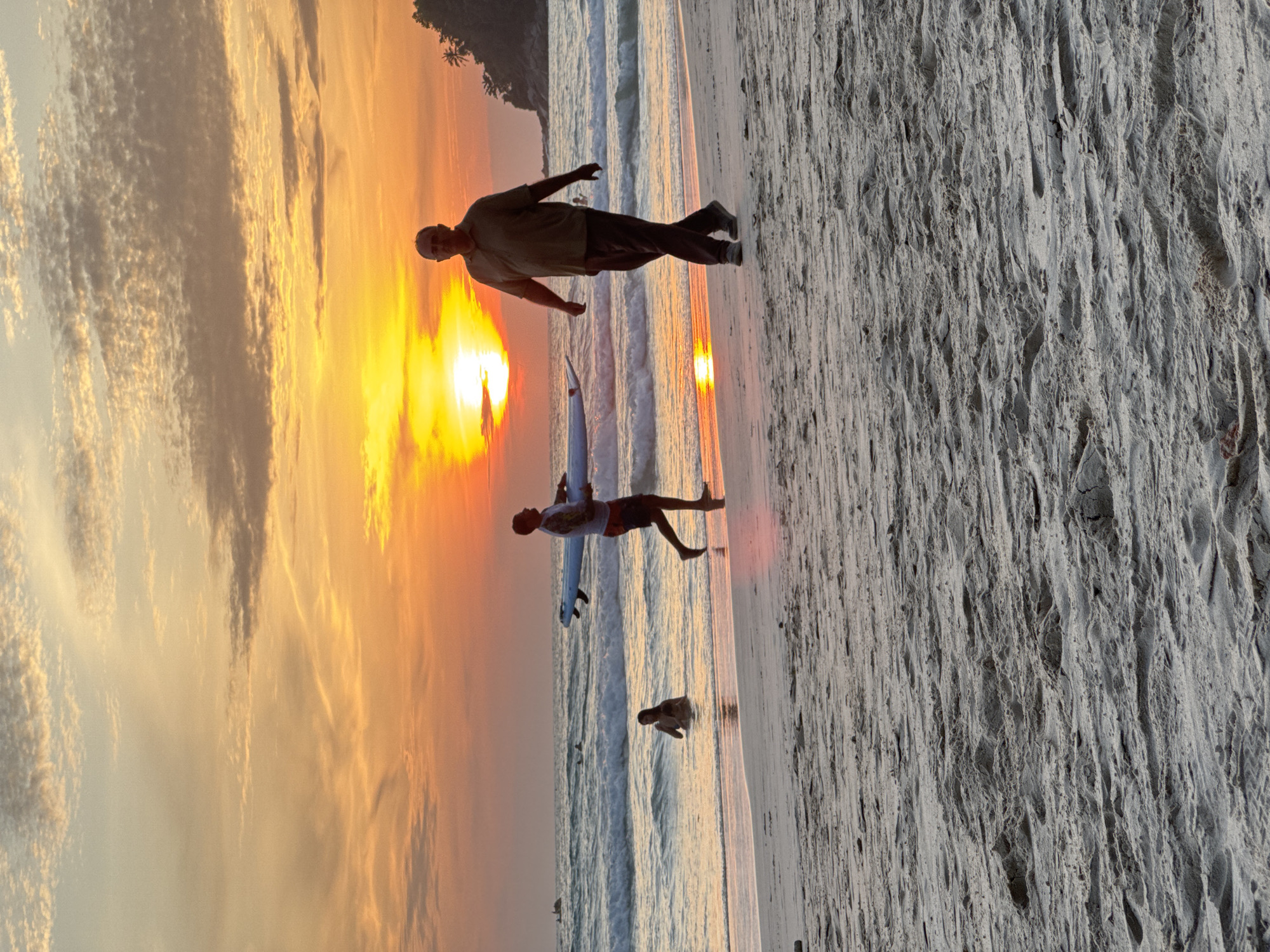 This image captures a sunset scene on Karon Beach in Karon, Thailand. The sky is filled with warm orange and yellow hues, with scattered clouds tinged by the setting sun. The ocean waves are gently rolling onto the sandy shore.
In the foreground, the sand is textured with footprints and ripples. In the middle ground, a person is walking on the beach carrying a white surfboard. To their left, a person is wading in the shallow water. Further out in the ocean, there are a few more figures visible. On the right side of the image, a tree-covered hill juts out into the sea, silhouetted against the vibrant sunset. The overall atmosphere is serene and tranquil.