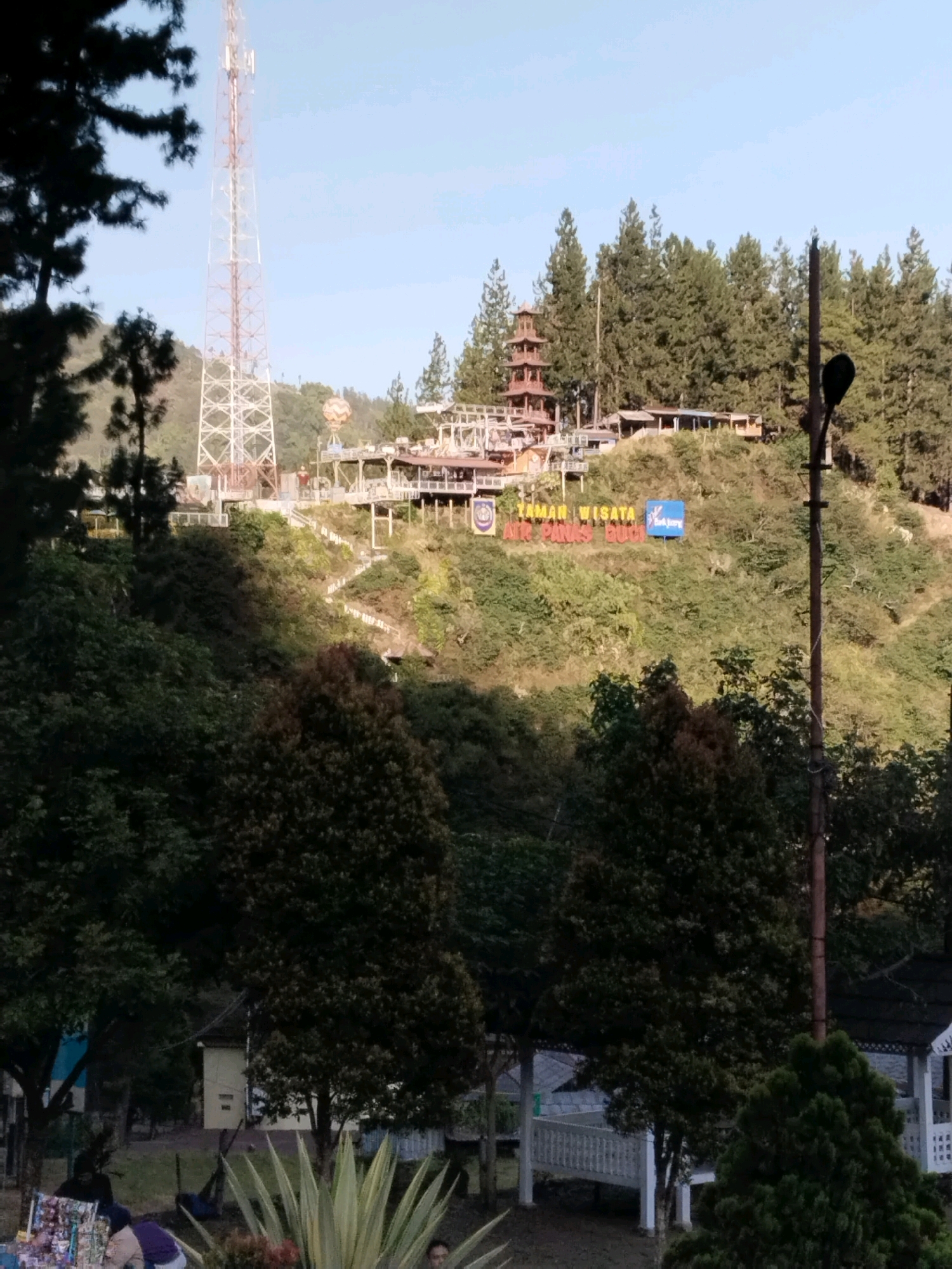 The image shows a wide view of a hillside tourist attraction in Guci, Indonesia, with buildings and a tall telecommunications tower on the upper part of the hill. The foreground is dominated by lush green trees and foliage. In the middle ground, a complex of structures, possibly hotels or restaurants, are visible on the terraced hillside, with a prominent pagoda-like building. A large sign reads "TAMAN WISATA AIR PANAS GUCI". A paved pathway zigzags up the hill towards the buildings. A tall, lattice-like telecommunications tower stands to the left of the main structures. In the lower part of the image, there are indications of a park or recreational area with some people gathered near stalls selling merchandise. The sky is clear and blue, suggesting a daytime scene.