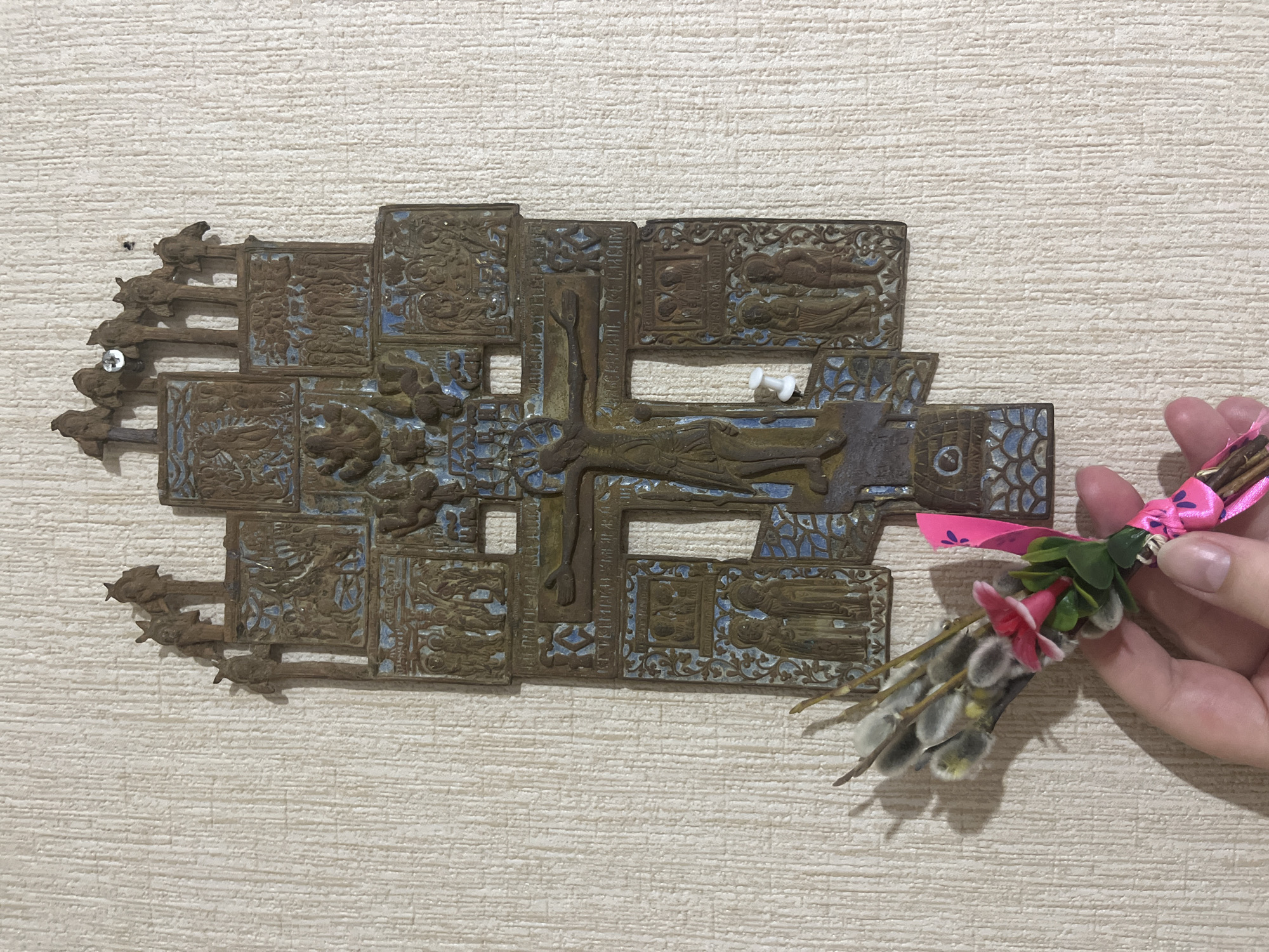 A tan textured wall serves as a backdrop for a bronze Orthodox crucifix and a hand holding a bouquet. The crucifix is intricately designed, with a central figure of Christ on the cross surrounded by rectangular segments depicting various biblical scenes and stylized ornamentation. The bronze surface shows signs of age and oxidation, with hints of blue paint or enamel filling some of the recessed areas. It is mounted on the wall with screws. Adjacent to the crucifix is a human hand holding a small bouquet of pussy willow branches, adorned with a pink ribbon, a small red flower, and green leaves. The location is Chaikovsky, Russia.
