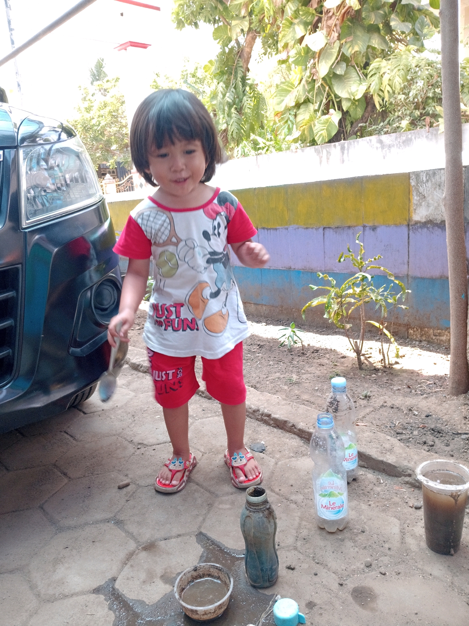 A young Indonesian child with short black hair is standing next to a dark-colored car in what appears to be a residential area in Gebang, Indonesia. The child is wearing a white t-shirt with a cartoon print and red shorts, along with red sandals adorned with cartoon characters. In their right hand, the child holds a metal spoon. To the right of the child, on the ground, are several plastic bottles, some containing water, and a dirty cup. The background features a textured wall with horizontal stripes of yellow and purple, overgrown with green foliage. The ground is a mix of dirt and paving stones. The scene is illuminated by natural daylight.