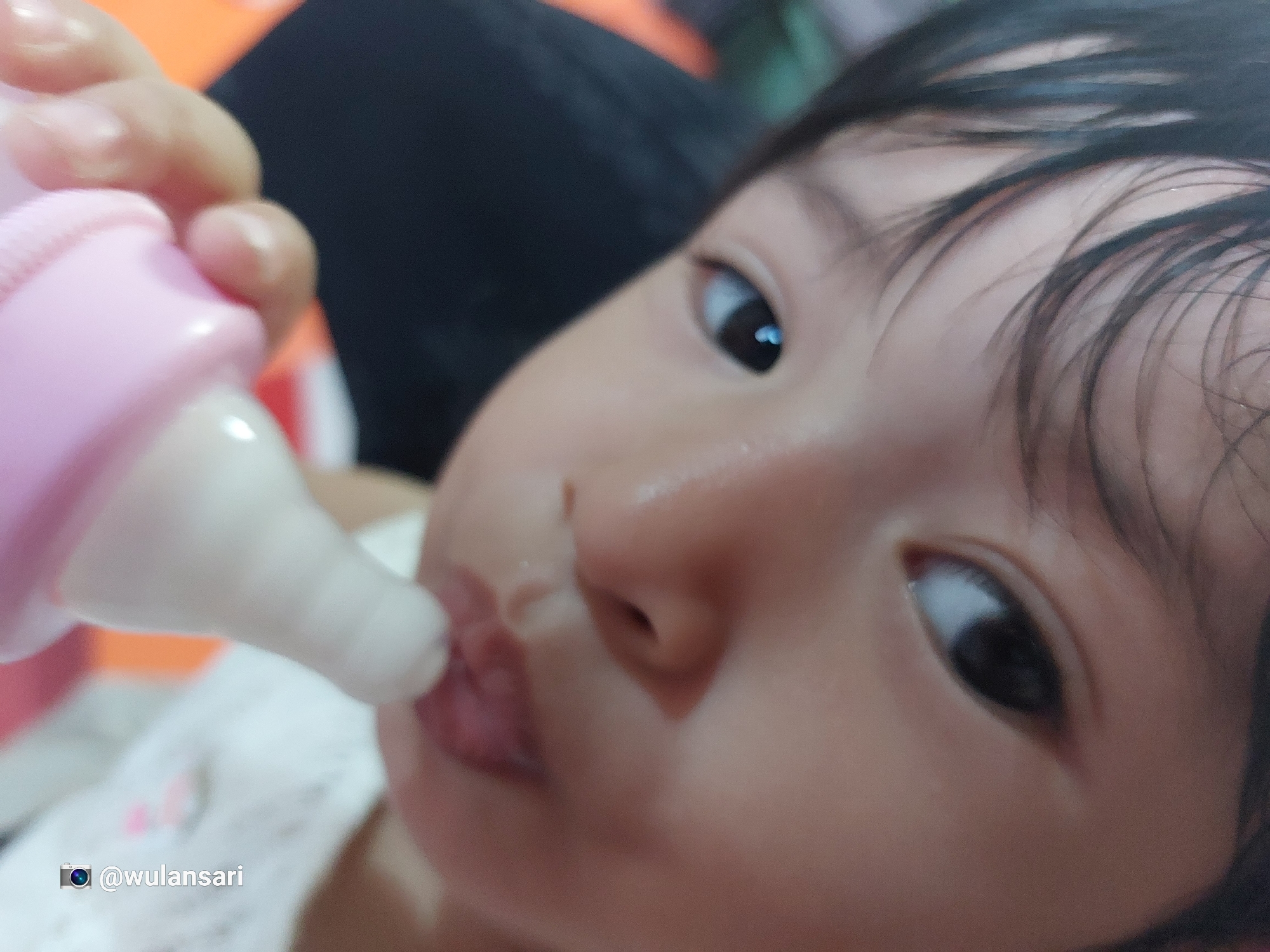 A close-up, low-angle shot shows a baby drinking from a pink and white bottle. The baby is fair-skinned with dark hair and dark eyes. The bottle is being held by a hand, likely an adult, partially visible in the frame. The background is out of focus and includes orange, black, and white colors. The photo includes the Instagram handle "@wulansari." The photo was likely taken in Meunasahblang, Indonesia.