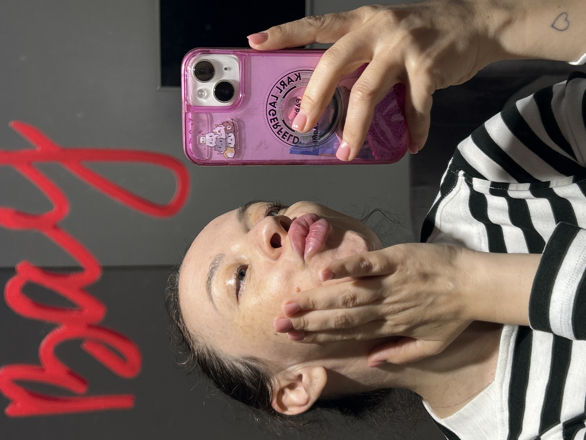 An upward-oriented mirror selfie captures a person holding a pink smartphone. The subject, visible from the chin upwards, has dark hair pulled back and wears a black and white striped long-sleeved top. Their face shows a playful expression, with eyes looking slightly upwards and lips pursed into an exaggerated pout. One hand covers the lower part of their face. The other hand holds a pink-cased smartphone featuring a dual camera system. The clear phone case is adorned with small cartoon characters and bears the reversed text "KARL LAGERFELD PARIS" around a circular design. A small, outline heart tattoo is visible on the wrist of the hand holding the phone. The background is a dark, reflective surface, with a blurry, red, cursive-like shape visible on the left, possibly decorative text like "Love." The lighting suggests an indoor setting.