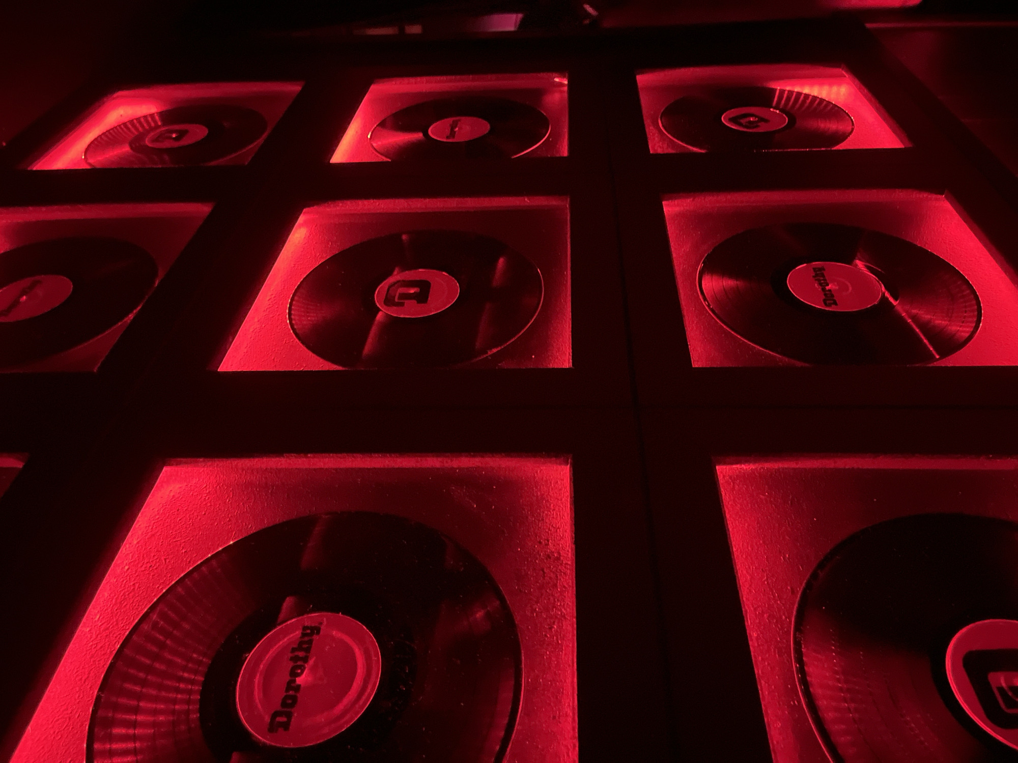 The image shows a grid of nine recessed squares, each containing a black record with a red label, illuminated by red light. The labels appear to be different, with one clearly reading "Dorothy". The scene is dimly lit, creating strong contrast and a sense of confinement. The location is Unknown, Unknown.