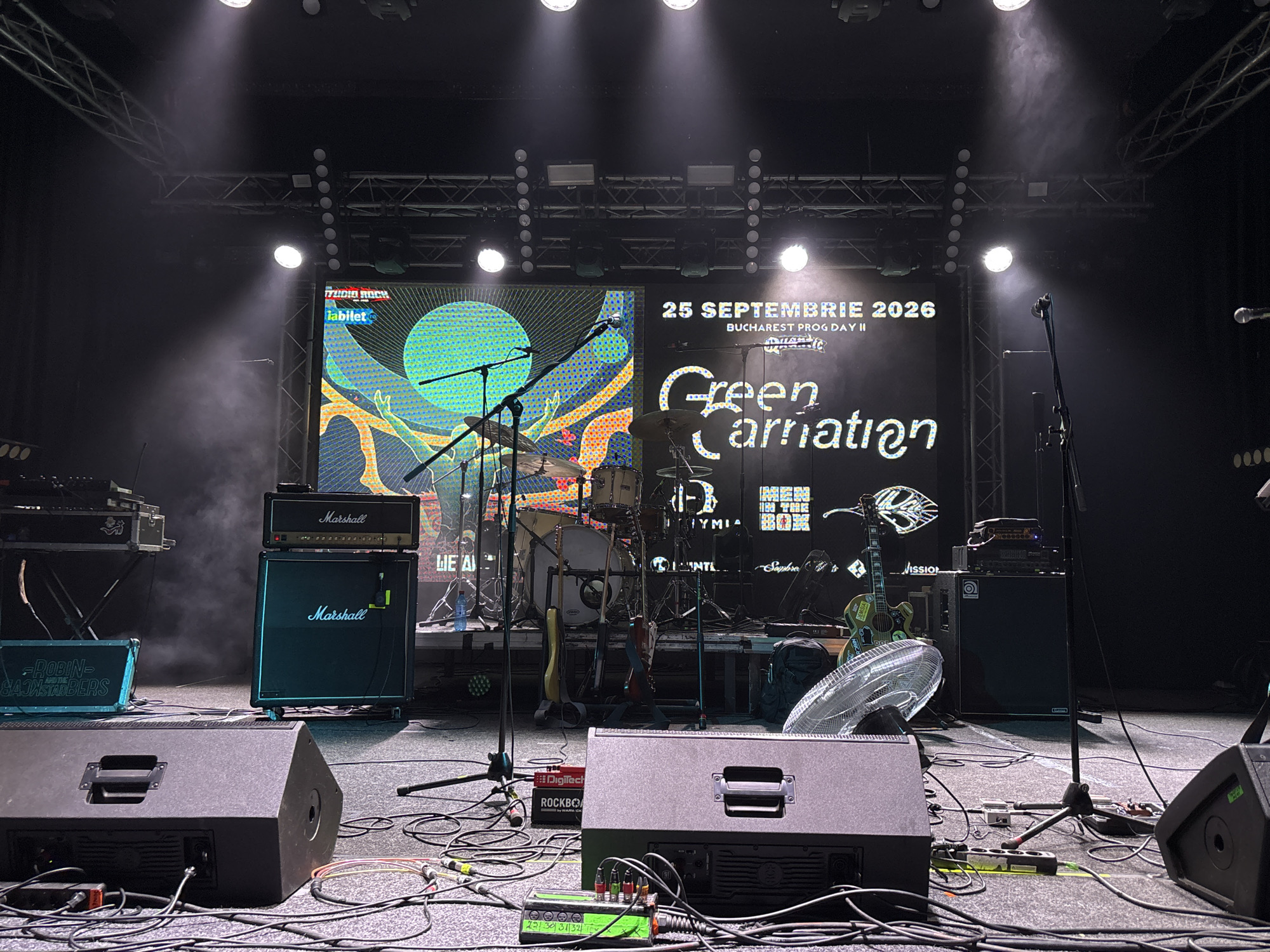 The image depicts a stage set up for a music performance, likely a rock concert, in Bucharest, Romania. The stage is equipped with musical instruments and audio equipment, including Marshall amplifiers, guitars, a drum set, microphones on stands, and large speakers. A large LED screen is prominently featured behind the stage, displaying graphics and text that identify the event as "25 SEPTEMBRIE 2026 BUCHAREST PROG DAY II" and featuring the band name "Green Carnation." The lighting suggests a concert atmosphere with spotlights creating beams of light and haze effects. The foreground shows a close-up view of audio cables and some of the stage equipment. The overall scene is well-lit and organized, indicating preparation for a live event.