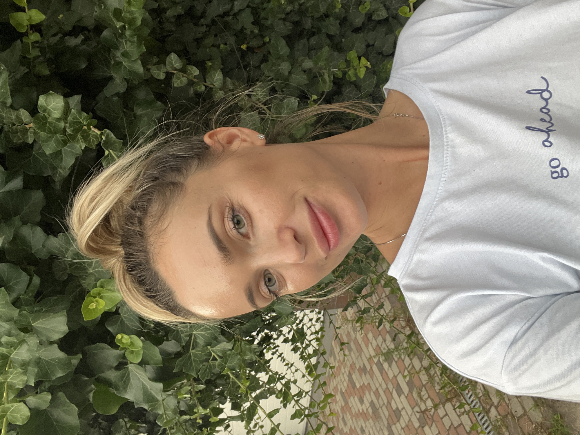 A blonde woman with her hair in a ponytail is taking a selfie in front of a wall covered in ivy. She is wearing a light blue t-shirt with the words "go ahead" written in dark blue cursive script on the chest. She has green eyes, light skin, and is smiling. To the left of the woman, a brick pathway is visible. The background consists of lush green ivy and a white wall. The scene appears to be outdoors, possibly in a garden or courtyard, in the city of Fontanka, Ukraine.