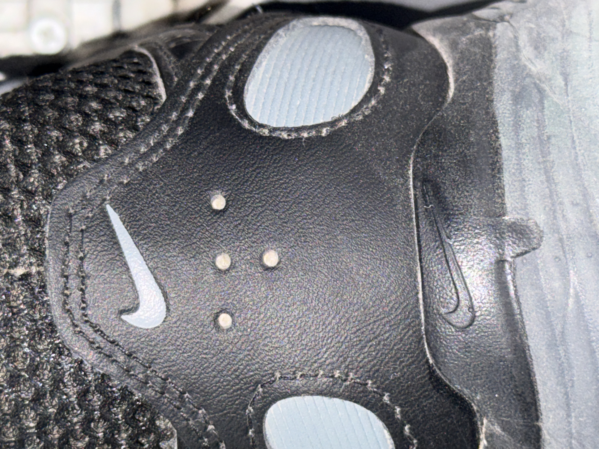The image shows a close-up, detailed view of the rear portion of a black Nike shoe. The shoe features a prominent white Nike swoosh logo on the heel tab. Below the swoosh, there is a debossed Nike logo on a textured black surface. The shoe has small, circular eyelets or decorative perforations on the heel counter, with three positioned centrally and two larger oval-shaped panels with a light blue ribbed interior on either side. The material of the shoe appears to be a combination of textured leather and mesh. The lighting suggests an indoor environment, possibly a studio or a room. There is no indication of the shoe's location in Cahul, Moldova.