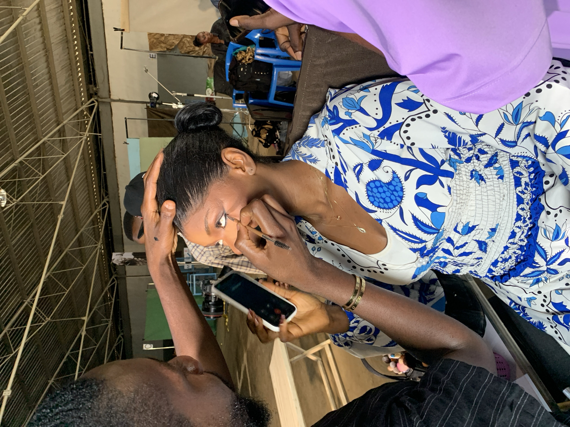 The image shows a close-up of a woman having her makeup applied. She has her hair pulled up in a bun and is wearing a white dress with a blue floral pattern. A makeup artist is applying eyeliner to her eye with a brush. The makeup artist is also holding a smartphone. In the background, there are other people and some studio equipment, suggesting that this is a professional setting, possibly a photoshoot or filming. The lighting appears to be artificial, indicating it could be indoors. The scene conveys a sense of preparation and attention to detail, focusing on the beauty process. The overall mood is calm and professional. The location is identified as Barakin Lerawi, Nigeria.