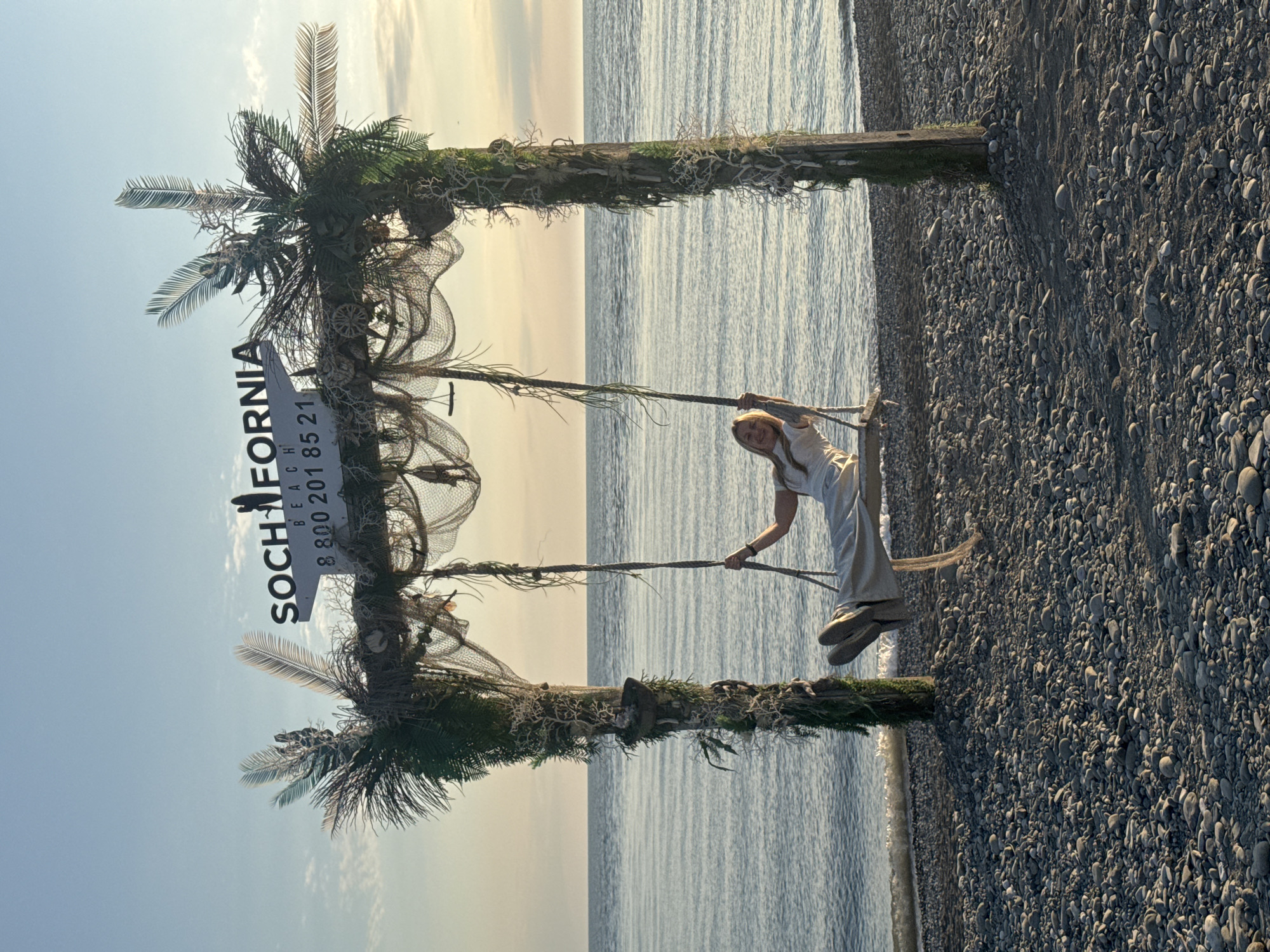 The image shows a woman standing on a swing in front of a decorative structure on a pebble beach. The structure consists of two wooden posts, decorated with artificial palm fronds, netting, and a sign reading "SOCHIfornia BEACH" along with a phone number. The background shows the sea stretching to the horizon under a sky with pastel colors suggesting dawn or dusk. The location is likely a beach in the city of Sochi, Russia.