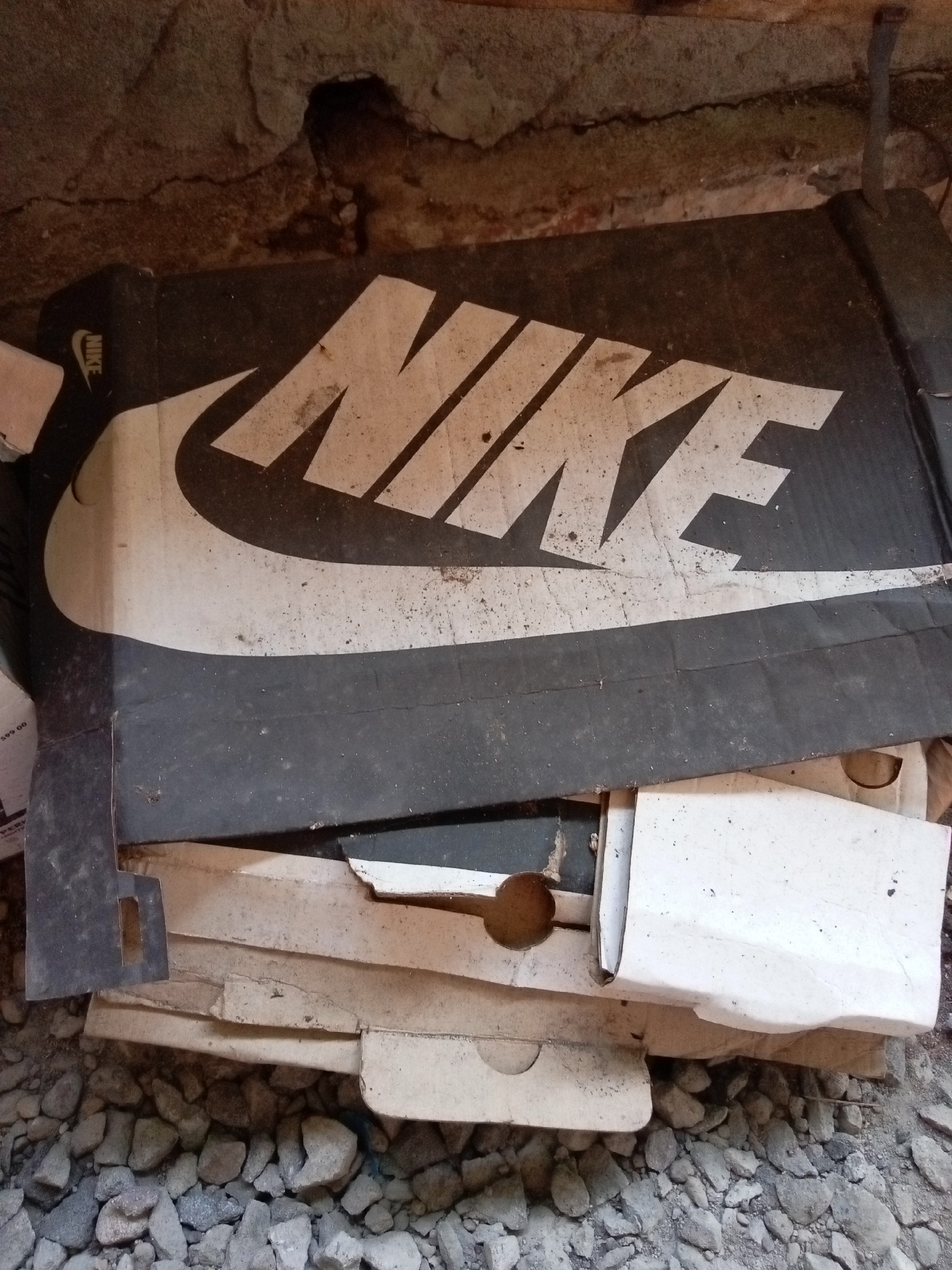 The image depicts a close-up, overhead view of a stack of cardboard boxes, predominantly a large Nike shoebox, lying on a surface of loose grey stones. The Nike box is black with the brand's white logo prominently displayed. Other cardboard boxes, some white and some with printed text, are partially visible beneath and around the Nike box. The scene appears to be outdoors, possibly in an area with a rough, unpaved floor, given the presence of the stones and the general appearance of disarray. The background consists of a textured, brownish surface, possibly a rough wall or foundation, with a small dark opening visible in the upper portion of the image. The lighting suggests it may be daytime, but the overall atmosphere is somewhat dim and dusty. There are no people or animals visible in the image. The exact location in Mojokerto, Indonesia, is not discernible from the visual information provided.