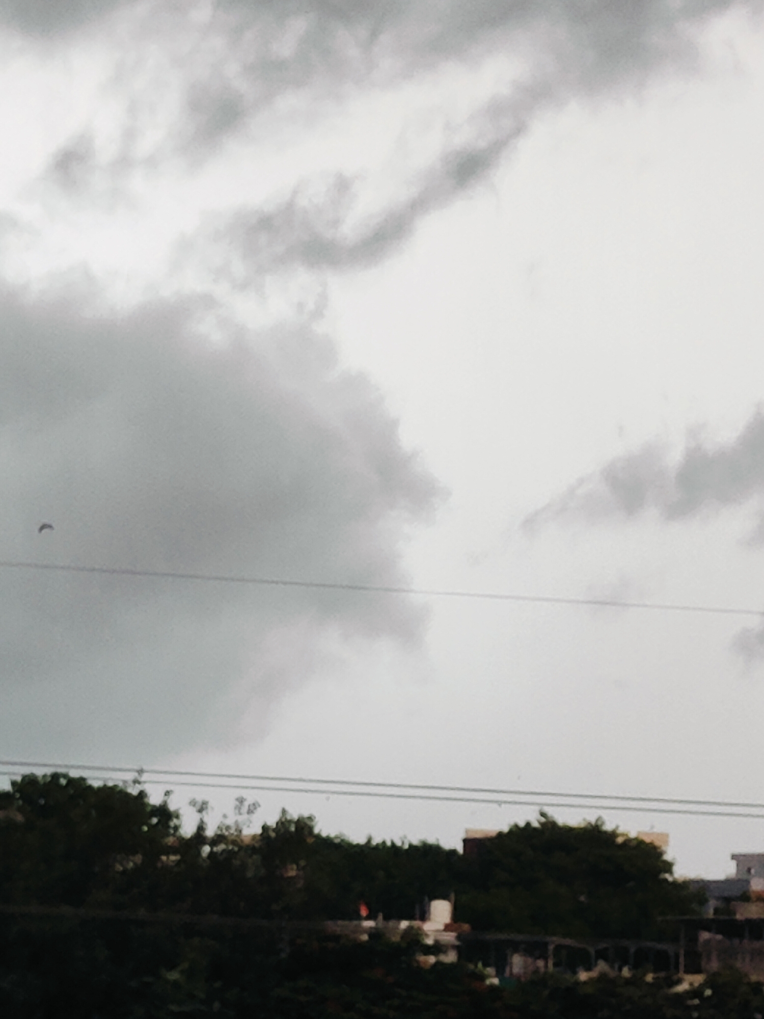 This is a low-angle, wide shot of a cloudy sky over Gurgaon, India. The clouds are thick and dark on the left side of the image, gradually becoming lighter and more diffuse towards the right. A thin, dark, curved line, possibly a wire or cable, is visible horizontally across the middle of the frame. In the lower portion of the image, the tops of trees are visible, obscuring the ground. These trees appear dense and dark green. Behind the trees, the tops of several buildings are visible, with flat roofs and light-colored walls. The overall impression is of an overcast day, possibly before or during rain.