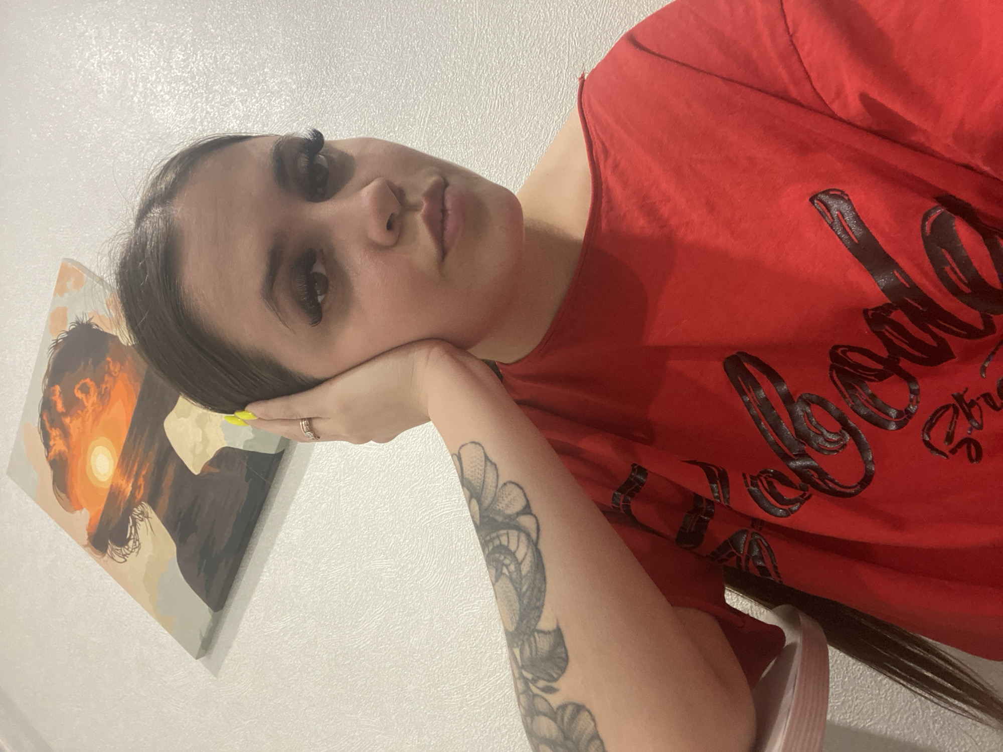 A woman with long brown hair, dark eyelashes, and light skin is resting her head on her left hand in an indoor setting. She is wearing a red t-shirt with black writing. A painting depicting a silhouette against a sunset is visible on the wall to her left. The woman has a tattoo on her left forearm. There is no indication that the image was taken in the city of Chaikovsky, Russia.
