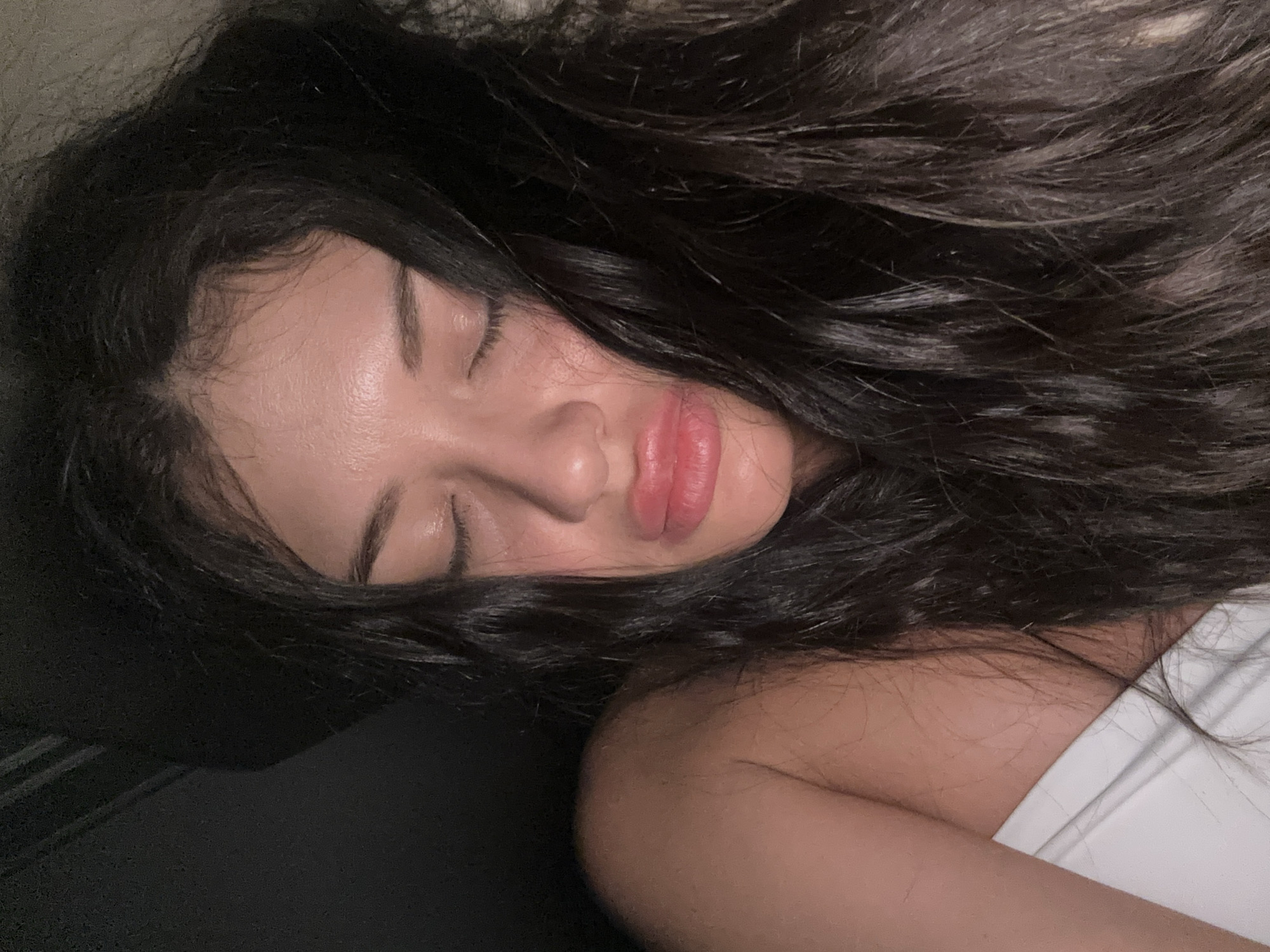 A close-up shot depicts a woman lying down with her eyes closed. She has long, dark hair that is spread around her head. Her lips appear full and slightly glossy. She is wearing a white top. The background is mostly dark, with what appears to be a dark surface behind her head. There is no indication of the location being Khimki, Russia, in the image.