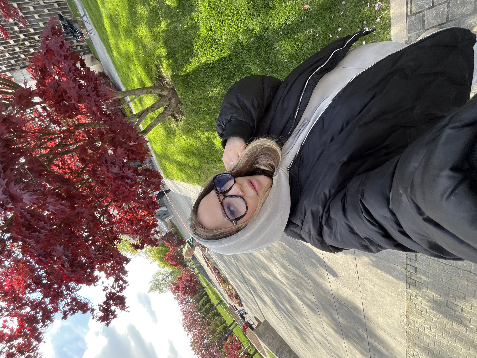 This image is a selfie taken outdoors by a young woman in what appears to be a park or public garden in Krasnodar, Russia.
The main subject is a young woman, likely in her early twenties, who is in the foreground, looking directly at the camera. She has blonde or light brown hair and is wearing dark-framed eyeglasses. Her attire consists of a black puffer jacket over a light grey hooded sweatshirt, with the hood up. A silver-colored ring is visible on the ring finger of her left hand, which is held near her face, suggesting she is holding the camera for the selfie.
The background features a vibrant natural setting with prominent red-leafed trees, likely Japanese maples, lining a paved path. Lush green grass patches are visible next to the path and beneath the trees. The path extends into the distance, with more red-leafed trees visible along its sides. The sky, partially visible, shows white clouds against a bright, clear blue background, indicating a sunny day. Shadows are cast on the path and grass, suggesting bright sunlight. Another blurry figure of a person can be seen further down the path in the background.
The activity taking place is the woman taking a selfie. The time of day appears to be daytime, likely morning or afternoon, given the bright lighting and visible shadows. The weather is sunny with some clouds. No visible text is present in the image.
