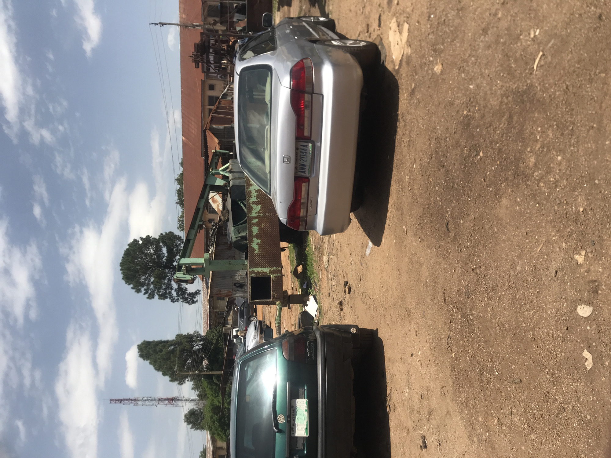 The image depicts an outdoor scene in Jos, Nigeria, likely a scrapyard or a workshop. Two cars are prominently featured: a dark green Volkswagen Golf in the foreground on the left and a silver Honda Accord on the right. The Golf appears to be older, with visible wear and tear, and is parked on a dirt lot. The Honda Accord, also older, is parked behind the Golf, facing away from the camera.
In the background, there are several structures with corrugated iron roofs, typical of the region. A tall, red and white radio tower stands to the left, and several trees are scattered throughout the scene. A large green industrial machine, possibly a crane or a loader, is also visible, adding to the industrial feel of the location. The ground is unpaved, consisting of dirt and gravel, with some debris scattered around. The sky is partly cloudy, suggesting a warm, sunny day. No people are clearly visible in the image, though there may be figures obscured by the vehicles or structures.