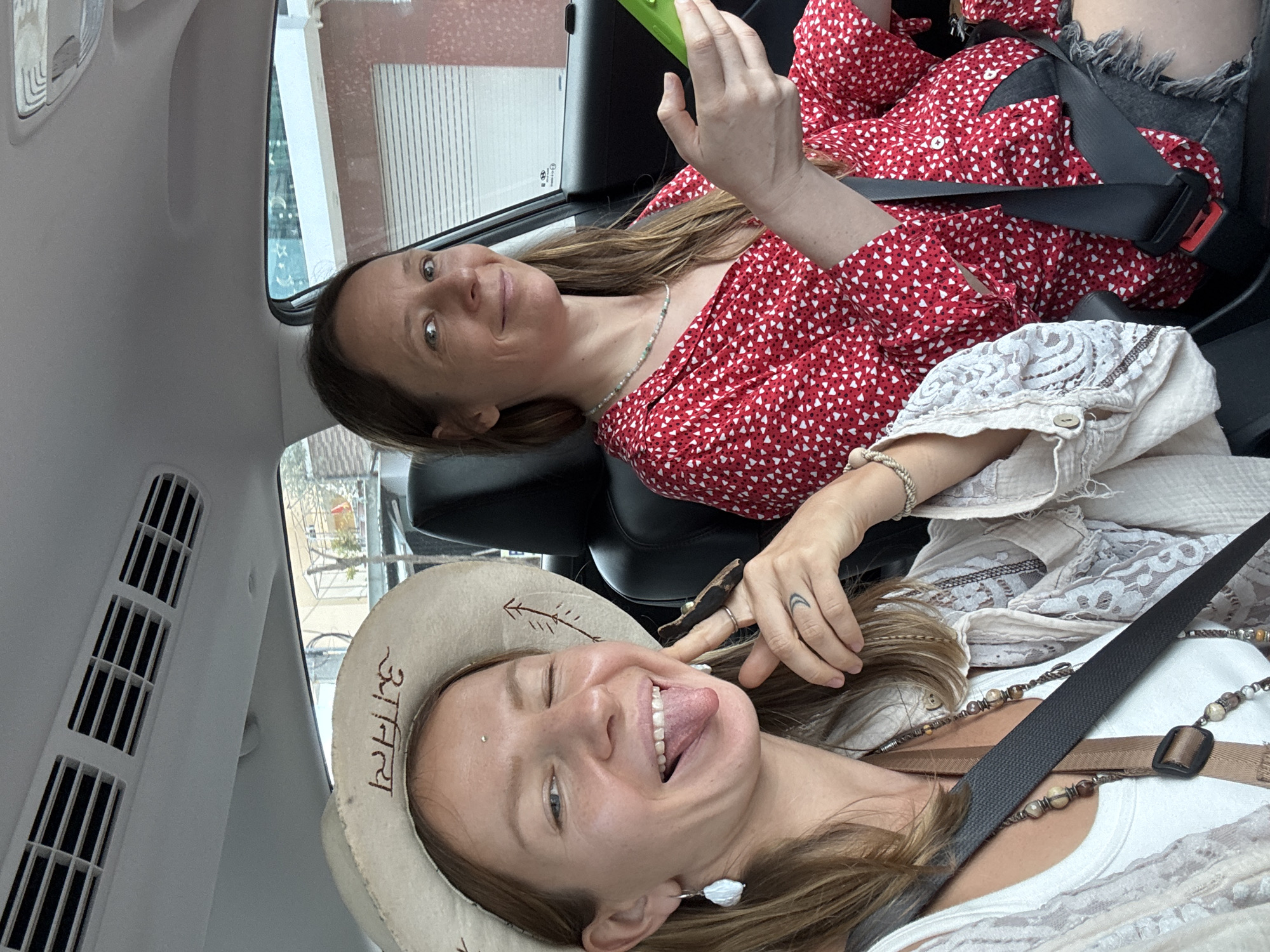 Two women are seated inside a vehicle, likely a car, based on the visible interior elements such as the dashboard, air vents, and seatbelts. The woman in the foreground, on the left, is wearing a light-colored cowboy hat and a white tank top with a light-colored lace overshirt. She is winking and sticking out her tongue, looking directly at the camera. She has a nose piercing and several rings on her fingers, one of which has a crescent moon tattoo on her hand. She is wearing a layered necklace. The woman in the background, on the right, is wearing a red shirt with a white heart pattern and dark shorts. She is looking towards the camera with a slight smile and holding a green object, possibly a phone, in her right hand. She is also wearing a beaded necklace. The vehicle appears to be in motion, as suggested by the slightly blurred background of buildings and a street scene which is consistent with an urban environment. The context of Phu Thuy Ward, Vietnam, is implied by external information and suggests an Asian urban setting. The overall mood of the image is casual and playful.