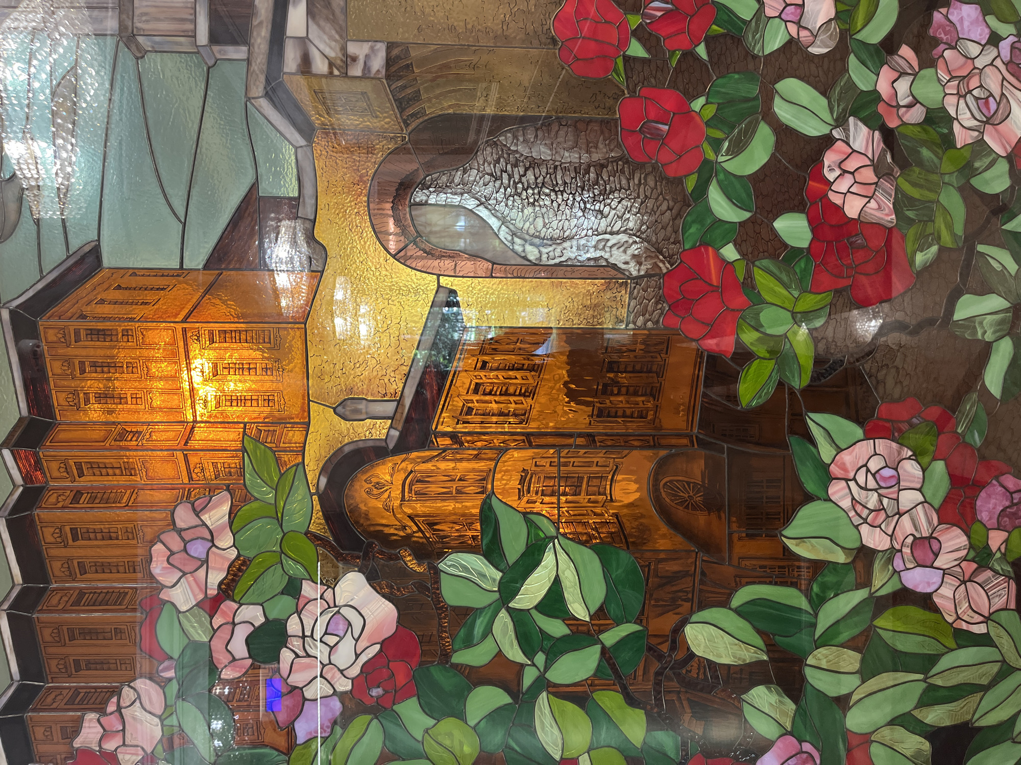 The image shows a stained-glass panel depicting a scene from Plovdiv, Bulgaria. It combines architectural elements and floral motifs. On the left, a building is depicted with multiple windows and levels. An arched structure reminiscent of an ancient aqueduct spans the central area. To the right, there are rose bushes with red and pink blossoms and green foliage. The architecture appears to be an abstracted cityscape.