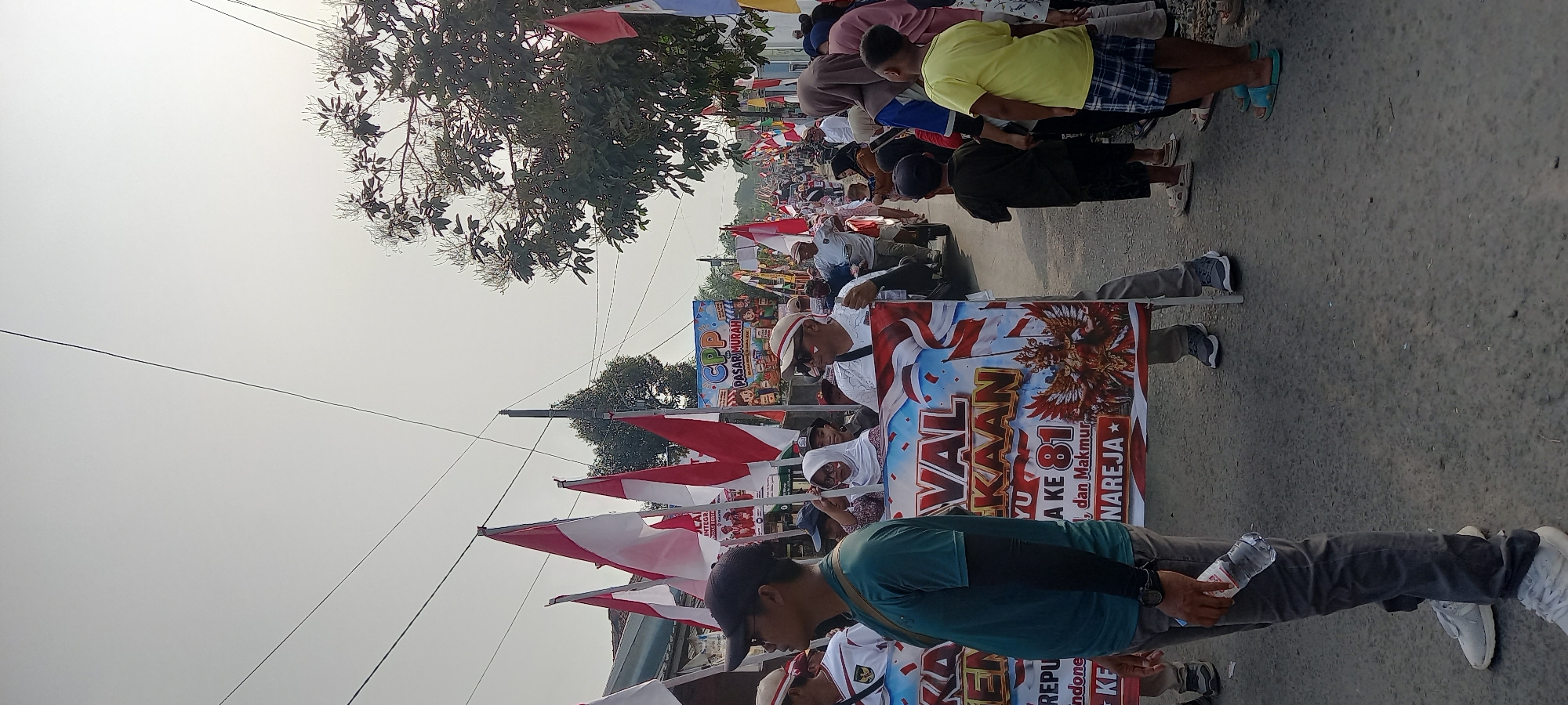 In Wanareja, Indonesia, a procession is underway on a paved street lined with buildings and trees. Several people are present, many holding or standing near Indonesian flags which are red and white. A large banner is prominently displayed, featuring the text "FESTIVAL KE-81 REPUBLIK INDONESIA" along with an image of Garuda Pancasila. Another banner in the background reads "CPP PASAR MURAH." Participants are dressed in casual attire, with some wearing hats and sunglasses. The sky is overcast, suggesting a daytime event. The scene depicts a celebratory atmosphere, likely a local festival or parade.