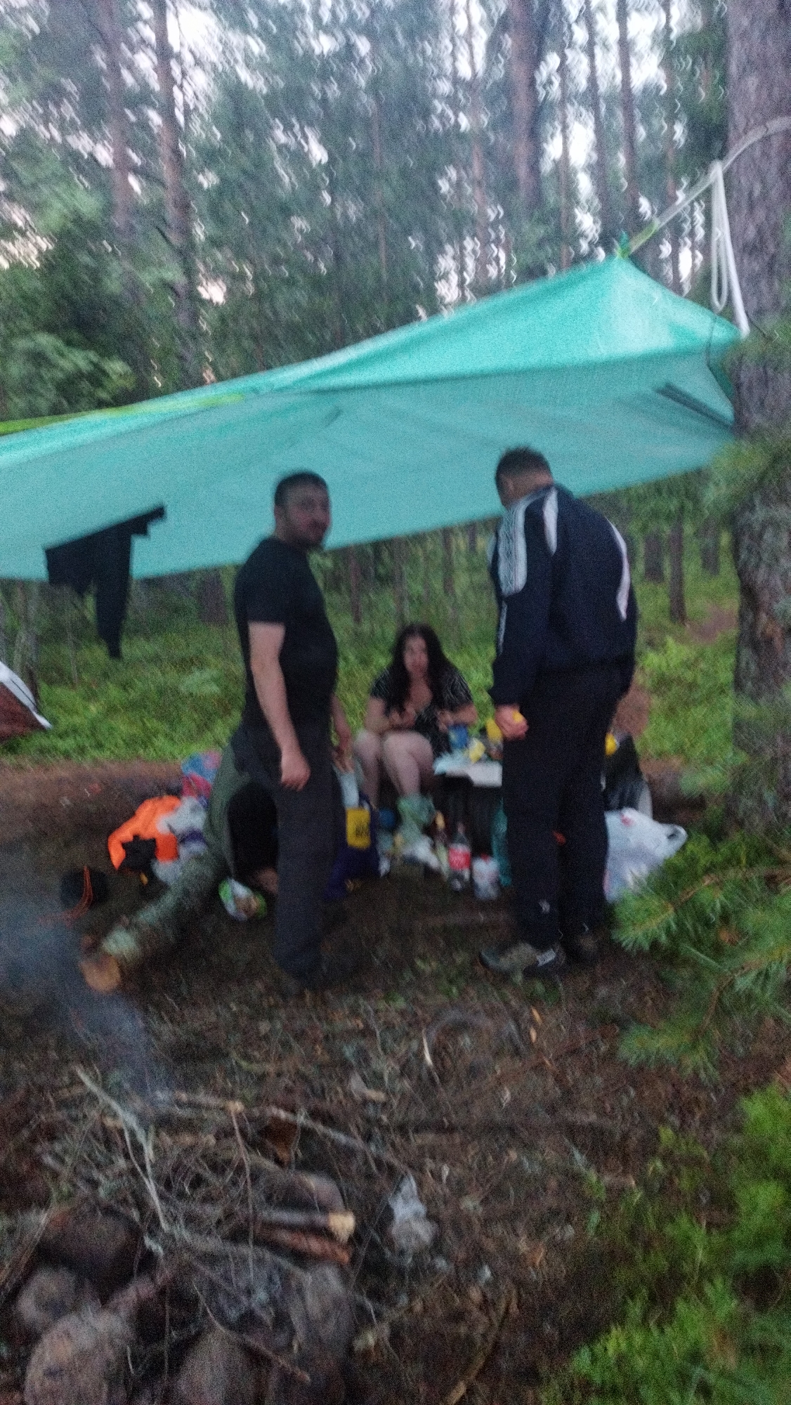 A group of four individuals is gathered in a wooded area in Tarasovskoe, Russia, under a teal-colored tarp shelter. Two men stand on the left and right of the frame, facing the center. The man on the left is wearing a black t-shirt and dark pants. The man on the right is wearing a dark jacket with white stripes and dark pants. A woman is seated in the center of the frame, wearing a black and white patterned dress. She is surrounded by various items including bottles, bags, and what appears to be food. The ground is covered with fallen leaves and branches, and a fire pit is visible in the foreground with smoking embers. The environment is a forest with tall trees and lush green foliage.