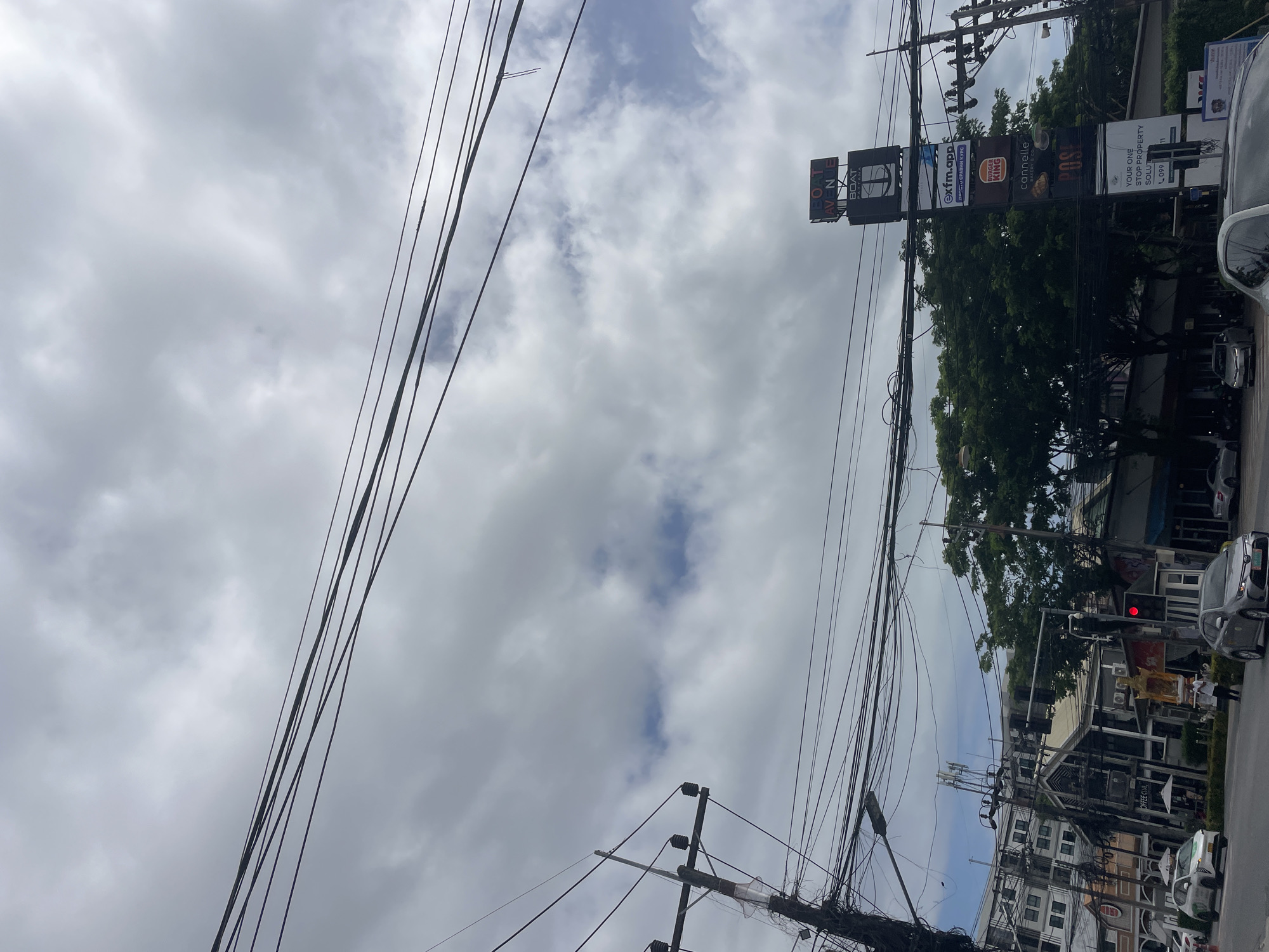 This image captures a street scene in Choeng Thale, Thailand, specifically at the Boat Avenue commercial area. The sky is largely overcast with white and grey clouds, though patches of blue sky are visible, particularly towards the upper right. A dense network of black electrical and communication wires crisscrosses the upper two-thirds of the frame, originating from and connecting to various utility poles, one of which is prominent on the left side of the image with multiple insulators.
The scene depicts a multi-lane asphalt road at the bottom, with several vehicles present. A white Toyota Yaris ATIV is positioned on the left, turning or stopped. Further right, two grey sedans are visible; one is in the foreground, and another is behind it. A red traffic light is illuminated on the right side of the road.
Along the street are various commercial buildings. On the left, there is a multi-story building with a distinctive facade and a sign partially visible that reads "COFFEE CLUB." To the right, a tall, vertical commercial sign dominates the mid-ground. From top to bottom, this sign displays "BOAT AVENUE," "BOAT PATTANA" (with an anchor logo), "exfm.app," the "BURGER KING" logo, "cannelle" (for Cannelle Bakery), "POSE" (partially visible), and text indicating "YOUR ONE STOP PROPERTY SOLUTION" with a phone number beginning "099." Lush green trees are visible behind and around this sign, providing a backdrop to the businesses.
One person is visible walking near the red traffic light, carrying what appears to be a tray with items. Another person is visible near the white car on the left. The overall impression is a bustling commercial area under a cloudy sky.