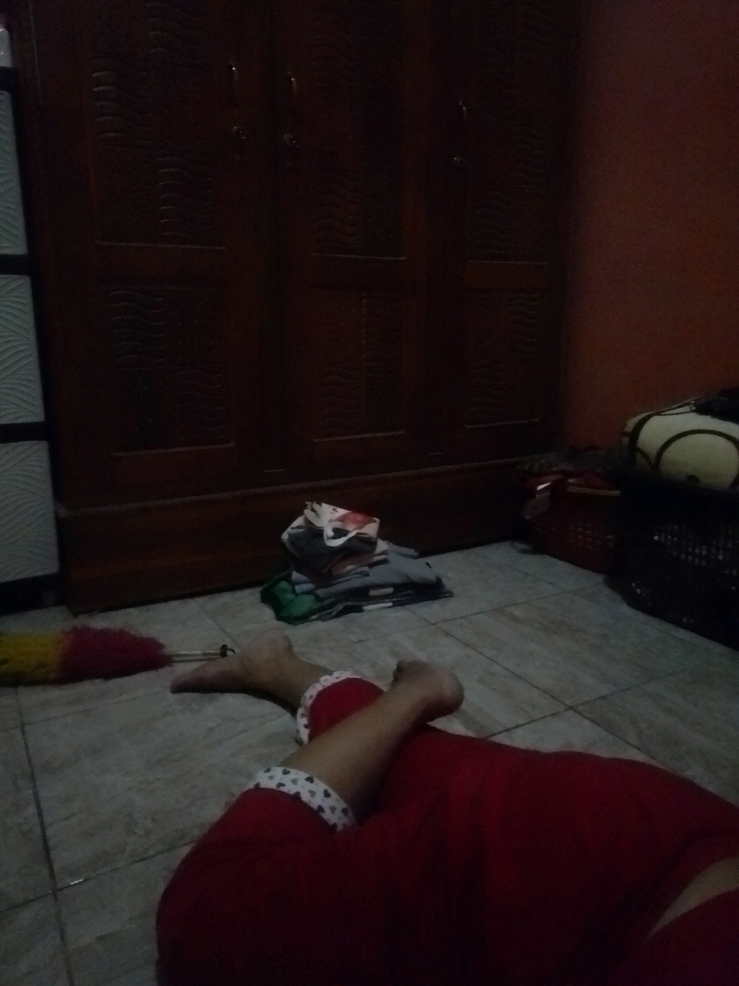 In a dimly lit room, possibly a bedroom in Kalijambe, Indonesia, a person is lying on the tiled floor. They are wearing red garments, with white trim adorned with small hearts around the ankles. A yellow and red feather duster lies near their feet. In the background, a large wooden wardrobe with carved doors stands against a wall. Nearby, a pile of clothes is stacked on the floor, and further to the right, there are baskets and what appears to be a decorative pillow with a black outline on a light background. The overall impression is one of a quiet domestic setting, with an emphasis on the personal space of the individual.
