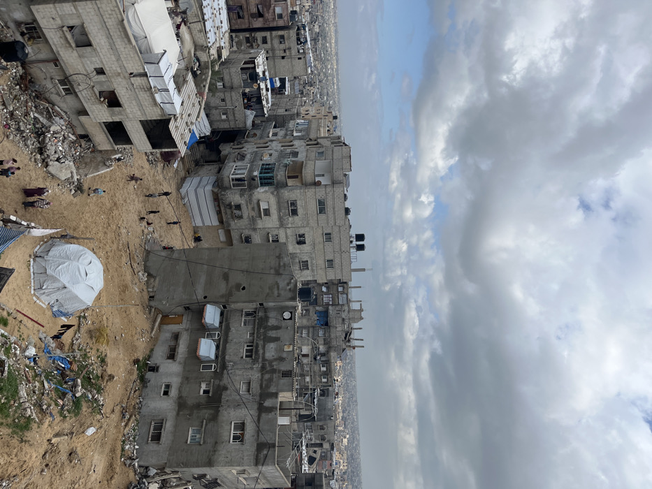 An elevated, wide-angle view depicts a densely constructed urban environment in Khan Yunis, Palestinian Territory, under an overcast sky with scattered blue patches. The scene is dominated by numerous multi-story concrete and cinder block buildings, ranging from three to six stories, exhibiting various stages of construction, repair, or damage. Many rooftops feature exposed rebar and black cylindrical water tanks.
In the foreground and mid-ground, significant areas of disturbed sandy earth and dirt separate the structures. Piles of concrete rubble and debris are evident, particularly in the lower left portion of the frame. A white, dome-shaped tent, consistent with temporary shelter, is positioned on the sandy ground in the mid-right foreground, surrounded by scattered refuse.
Approximately ten to fifteen individuals are visible on the sandy ground. A group of children appears to be congregated and active in the central dirt area, suggesting play. Closer to the white tent, a smaller group of adults, some wearing head coverings, stands on the sandy terrain. The distant horizon reveals a continuation of the urban sprawl, characterized by similar building types.