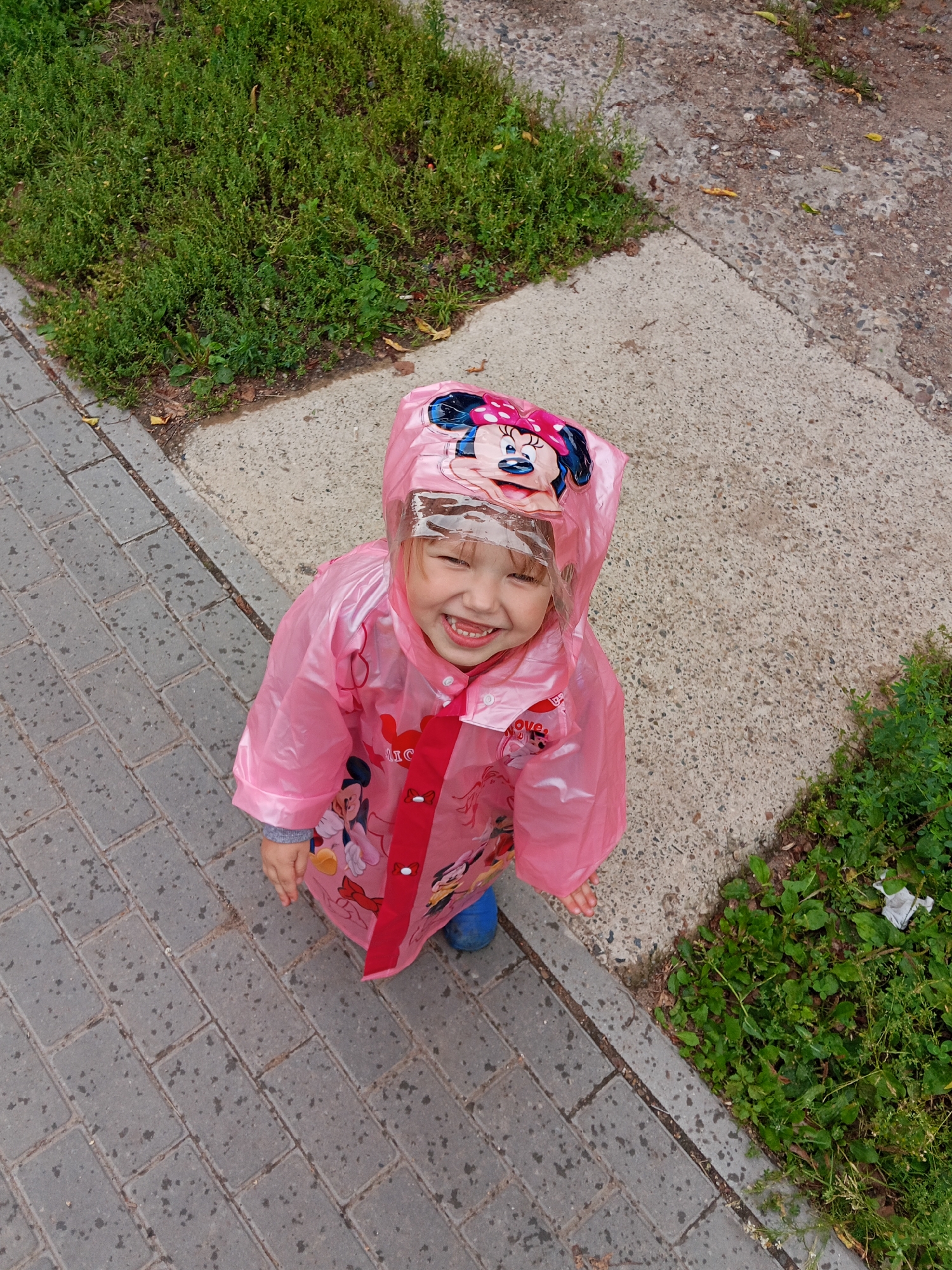 A young child, wearing a pink Minnie Mouse-themed raincoat and blue boots, is smiling and looking up at the camera. The child is standing on a wet paved walkway, next to a patch of green grass and a concrete area. The background shows more grass and a dirt path. The scene appears to be outdoors in an urban environment.