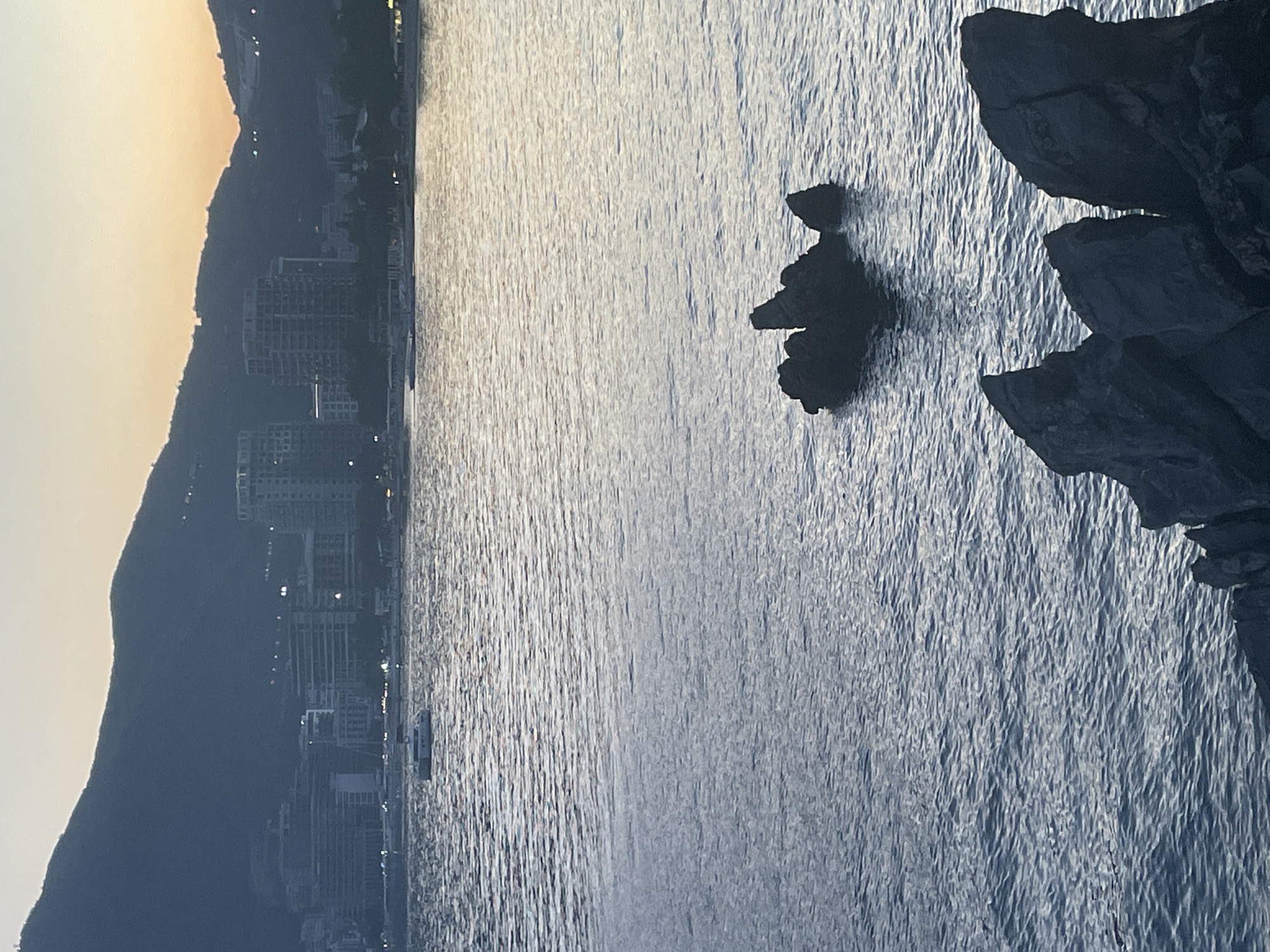 The image captures the coastal scenery of Budva, Montenegro, with the Adriatic Sea dominating the view. The water's surface exhibits subtle textures, likely caused by wind or currents. In the midground, several rocky outcrops emerge from the sea. The shoreline features a silhouette of a mountain range and a cityscape with buildings. The sky is visible at the upper left, with a pale gradient suggesting either sunrise or sunset. The rocky features in the foreground appear to be part of a larger cliff formation.