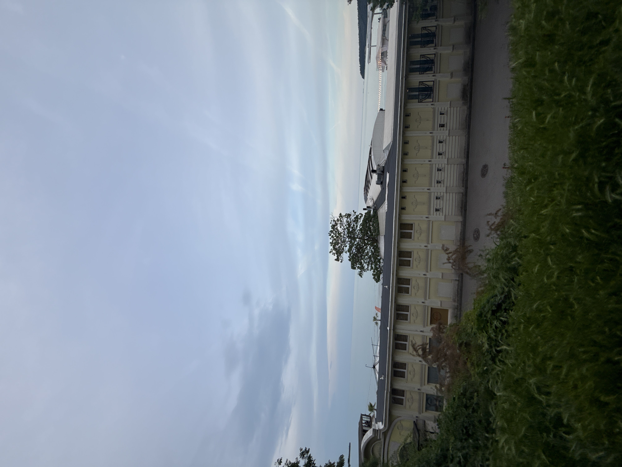 The image depicts a scene in Varna, Bulgaria. A multi-story building with light-yellow walls and symmetrical window patterns occupies the right side of the frame, bordered by greenery. A single tree stands near the building’s edge. Beyond the building and tree lies a body of water, likely the Black Sea, with a visible shoreline in the distance. The sky is overcast with varying shades of gray and light blue, creating a muted atmosphere. There are no people or apparent interactions visible in the image. A walkway is visible next to the building.