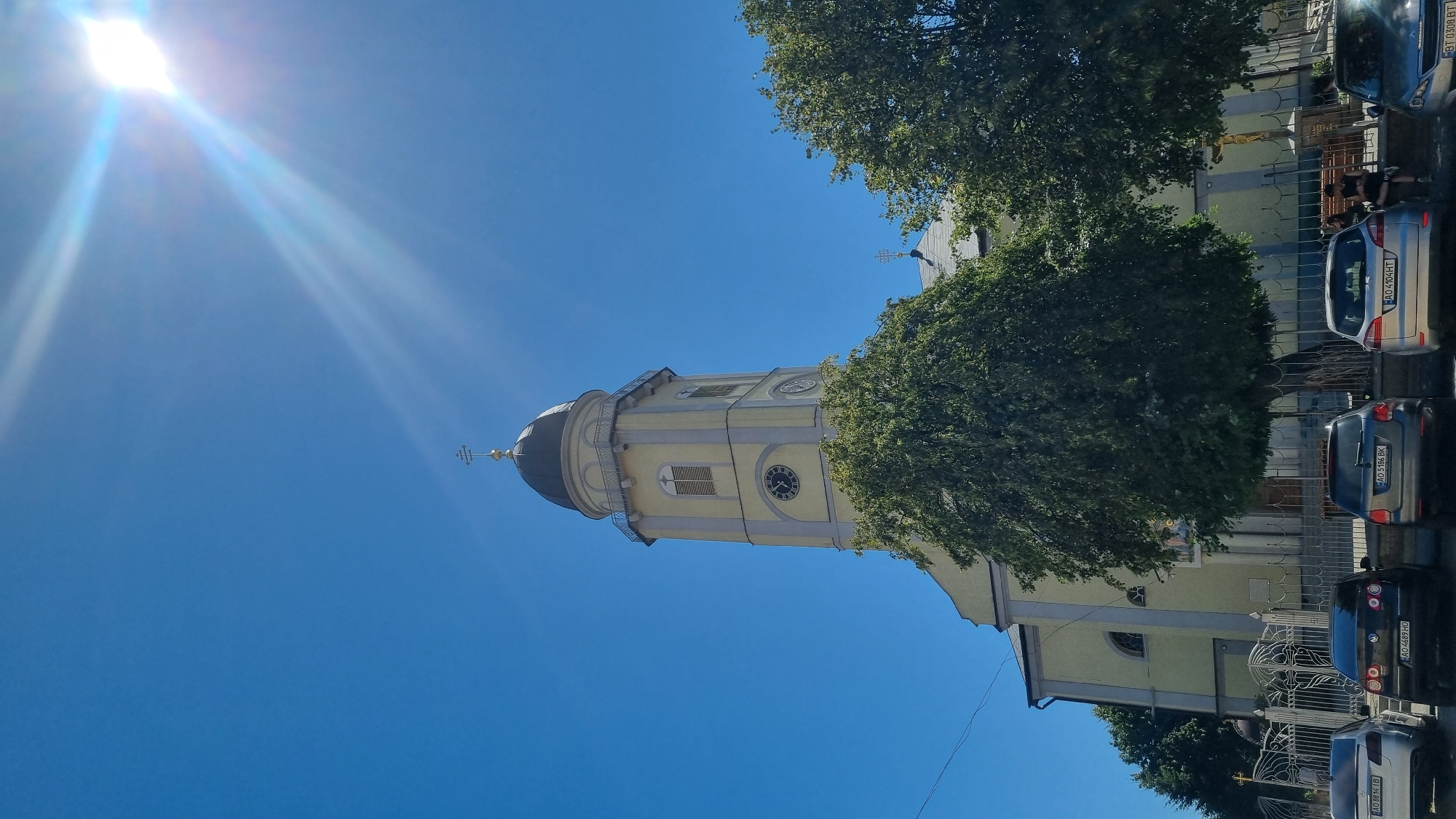 This is a daytime, outdoor, low-angle photograph of a church in Mukachevo, Ukraine. The church features a tall, pale yellow tower with a dark dome topped by a cross. The tower has visible clock faces and arched windows. A large, leafy green tree stands in front of the church, obscuring part of its facade. In the foreground, several cars are parked, and a metal fence runs along the perimeter. The sky is a clear, bright blue, with the sun prominently visible in the upper right corner, casting lens flare across the image. Two people are seated on a bench near the fence, on the right side of the frame. The overall scene depicts a sunny day in a city.