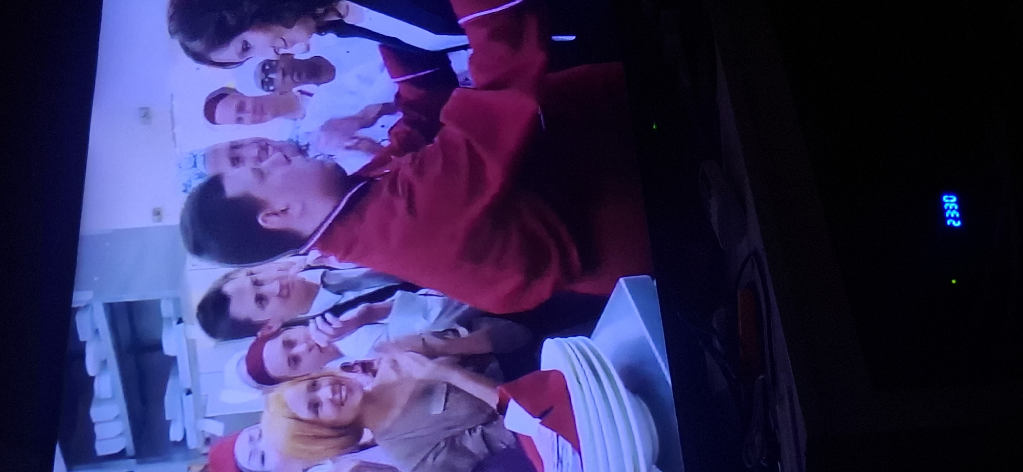 The image shows a dimly lit television screen displaying a scene with multiple people. In the foreground, a man wearing a red jacket is embracing a woman. To the left of the man, a woman with short blonde hair is smiling and clapping her hands. Behind them, several other individuals are visible, some wearing uniforms or formal attire. In the background, shelves stocked with white items, possibly plates or food containers, are visible. The environment appears to be indoors, possibly a catering or event setting. The lower right corner of the screen shows a blue digital display reading "2330" and a green illuminated dot. The scene is captured from an angle, suggesting it is a photograph of a television. The specific location in Yerocham, Israel, cannot be determined from the visual information provided in the image.