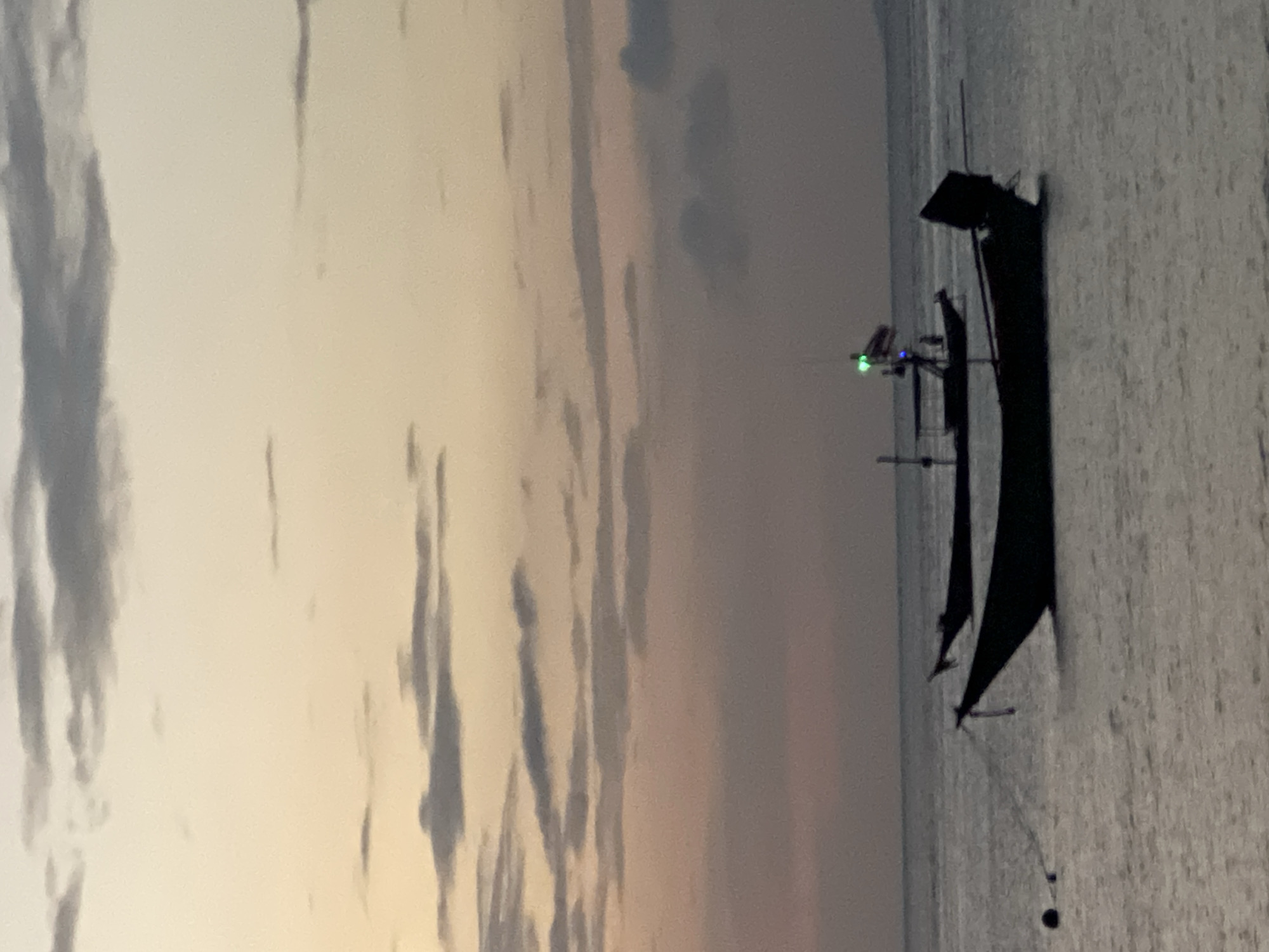 The image depicts a serene ocean scene at dusk in the Ko Pha-ngan District, Thailand. Two silhouetted fishing boats are anchored in the calm water, their dark forms contrasting with the soft hues of the twilight sky. The sky is a gradient of pale yellow, pink, and gray, with wispy clouds scattered across it, indicating the transition from day to night. The water is still, reflecting the muted colors of the sky. The foreground boat is closer to the viewer, showing a mooring line with a buoy. The second boat, further away, has a flagpole with a flag and a green light visible at its stern. The overall atmosphere is peaceful and tranquil, typical of a coastal sunset.