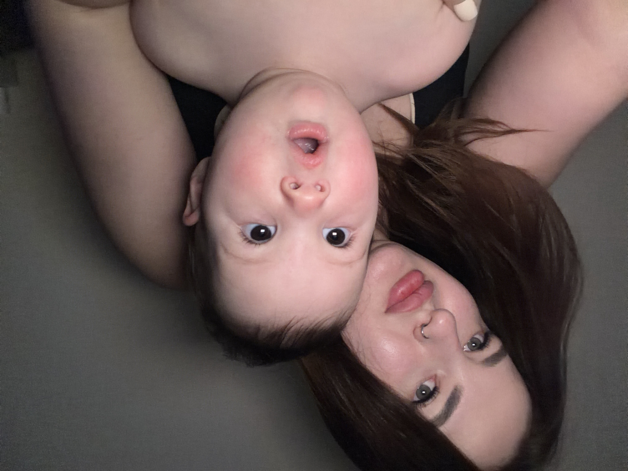 The image depicts an adult female and a baby. The baby is positioned above the female, with its face visible. The adult female's face is visible, with her cheek touching the baby's head. The baby has an open mouth. Both the female and the baby are fair-skinned. The female has dark brown hair. The location is not identifiable as Krasnodar, Russia.