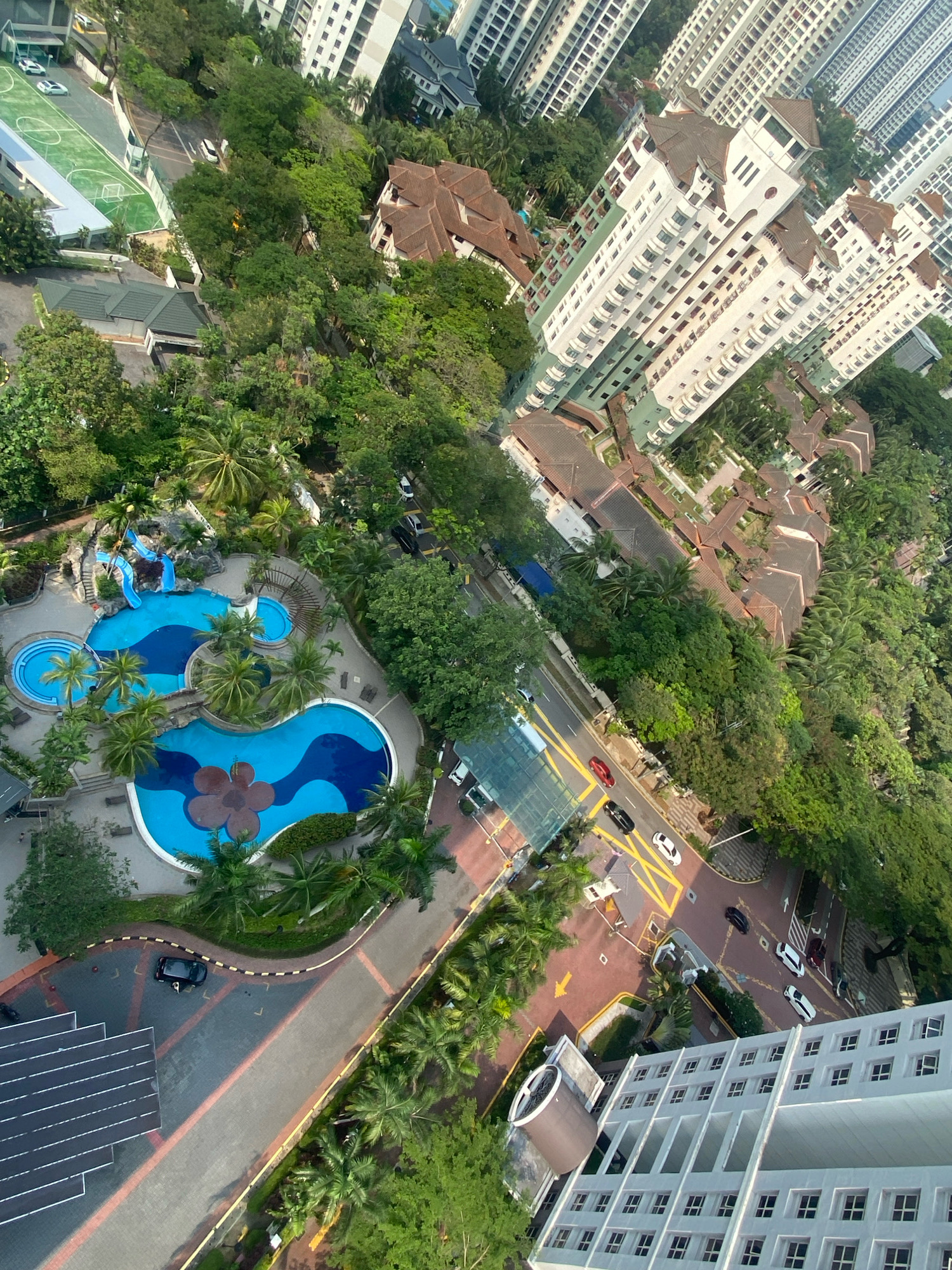 This is an aerial photograph of a residential complex in Kuala Lumpur, Malaysia, likely in a tropical urban setting. The scene showcases a resort-style swimming pool with blue water and a flower-shaped design in the foreground, surrounded by lush greenery and palm trees. Adjacent to the pool area is a multi-story apartment building with a light-colored facade and numerous windows. In the background, several taller, modern apartment buildings with white facades and many windows are visible, indicating a dense urban environment. The photograph also shows roads with vehicles, a sports court with green turf and markings, and a series of low-rise buildings with brown tiled roofs, suggesting a well-developed residential area. The overall impression is one of a large, well-maintained housing development with amenities integrated into a tropical landscape.