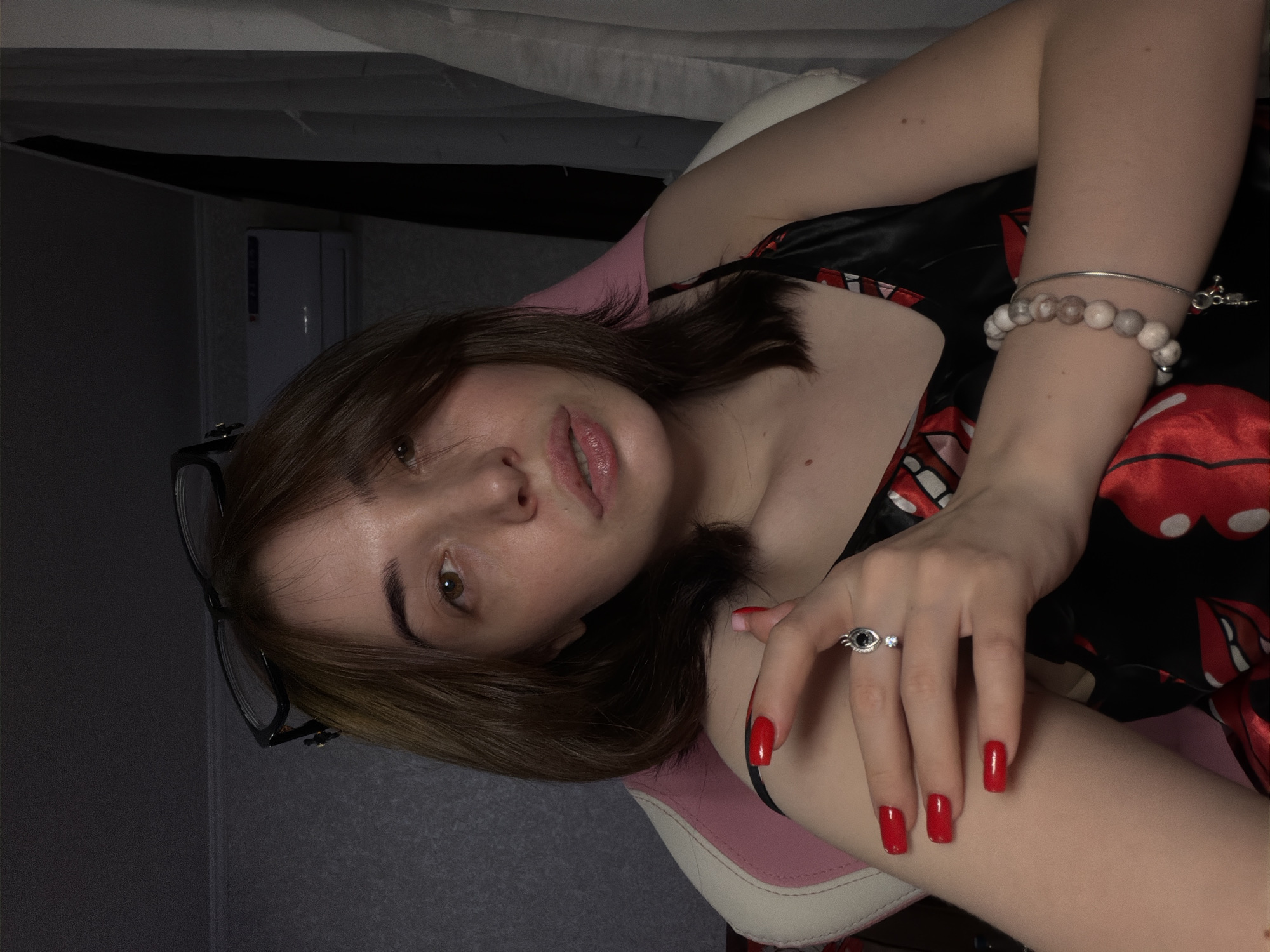 A woman with brown hair, wearing a black and red silk camisole, sits in a pink and white gaming chair. She has glasses perched on her head, bright red nail polish on her fingernails, and a ring with a black stone on her left hand. A bracelet adorns her right wrist. The background shows a grey textured wall, a white curtain, and an air conditioning unit mounted high on the wall. The lighting is dim, casting shadows.
