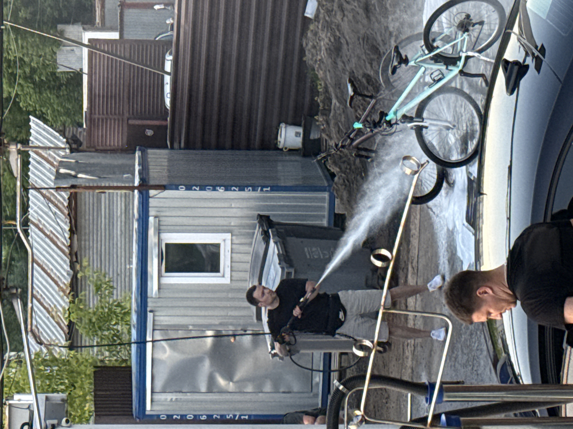 In Moscow, Russia, on a cloudy day, a man in a black t-shirt and grey shorts is pressure washing two bicycles at a car wash. The bicycles are light blue and dark red. The man is holding a pressure washer and spraying water on the bikes. In the background, there is a grey metal shed, a large grey trash bin, and a fence. The ground is covered in dirt and water. In the foreground, there is a black car and part of a person's head is visible. The scene suggests a casual outdoor activity.