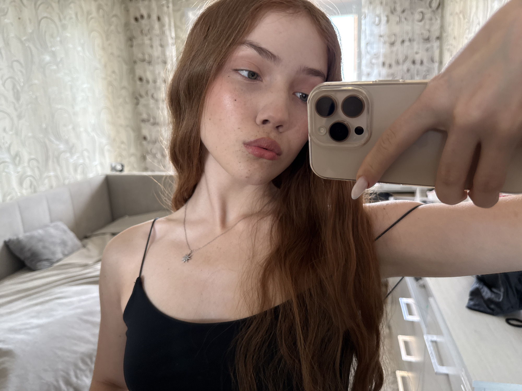 A young woman captures a mirror selfie in what appears to be a bedroom or private living space in Naberezhnye Chelny, Russia. She holds a light-colored iPhone with a triple camera system, partially obscuring her face. The woman has long, wavy reddish-brown hair, fair skin dotted with noticeable freckles, and light-colored eyes. She wears a black spaghetti-strap camisole and a delicate silver necklace featuring a star-shaped pendant. Her left hand, holding the phone, displays manicured nails. Her head is tilted slightly, and her lips are pursed in a subtle pout, with her gaze directed somewhat to the side, reflecting her pose for the selfie.
The background shows a neatly made bed with a grey headboard and a matching throw pillow. Behind her, light-colored curtains with a subtle scroll pattern hang, suggesting natural light is entering the room, indicating it's daytime. Modern, light-toned furniture, possibly a dresser or vanity, is also visible. The overall ambiance is bright and contemporary. There is no visible text in the image.