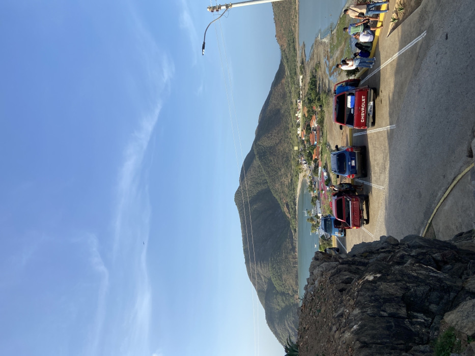 A steep hillside view reveals a road lined with vehicles, some red and one blue, including a red Chevrolet pickup truck. People gather near the road's edge. Below, a small town nestles along a lake's shore, framed by a large, dry mountain. A power line extends into the clear blue sky with a few wispy clouds. Rocks and foliage are visible in the lower foreground.