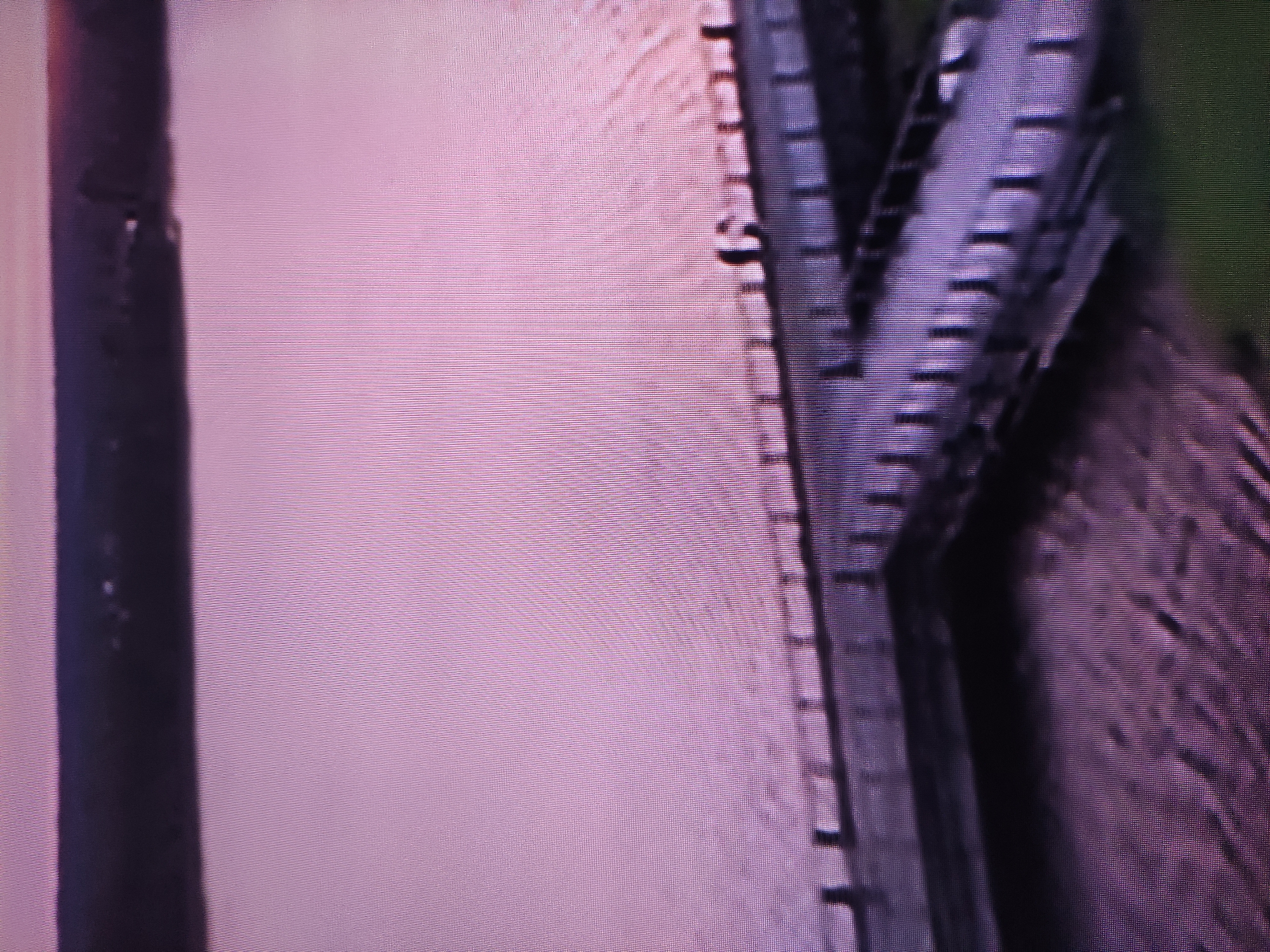 The image shows a close-up of metal structures, possibly a bridge or industrial equipment. The structures are dark, possibly painted a dark color, and feature repeating patterns of horizontal lines and vertical supports. To the left, a vertical pipe or post is visible. The background consists of a light-colored, flat surface with a uniform texture. The image is grainy and pixelated, suggesting it may be a low-resolution photograph or a detail from a larger image. There is no information that the image is taken in Pavlodar, Kazakhstan.