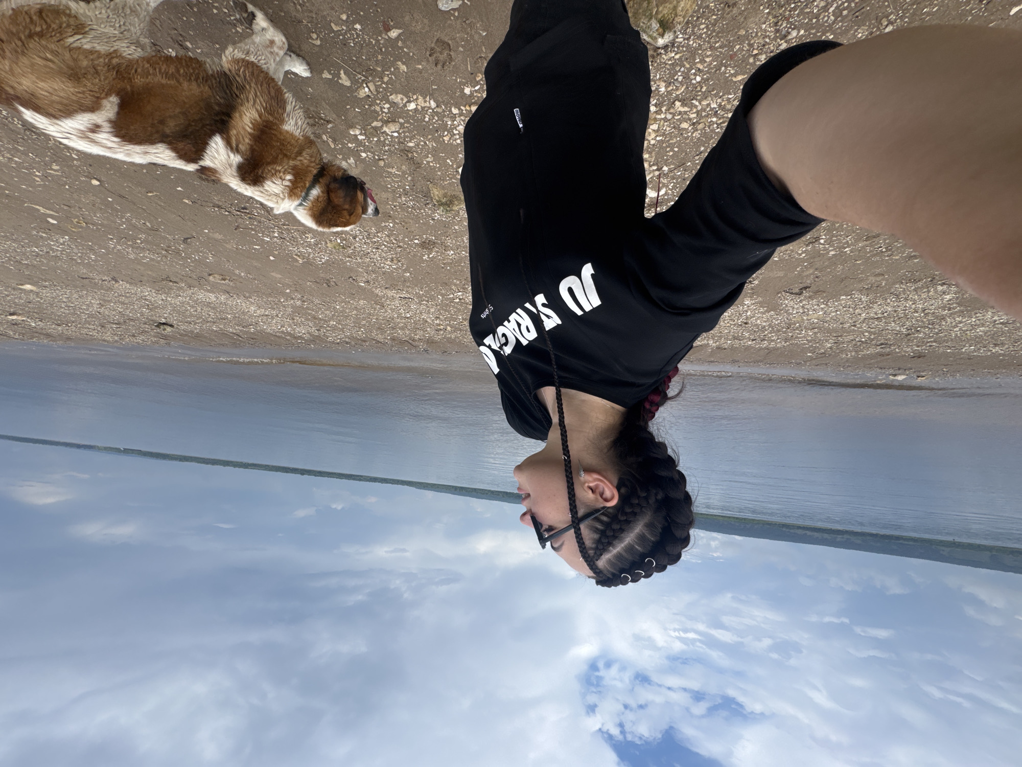 A young woman with braided hair styled with metal rings stands on a pebble-strewn beach near a body of water. She is wearing a black hoodie with the text "JUST RAGGA" printed in white on the front, and dark pants. She is holding up her left arm as if taking a selfie. To her right, a medium-sized dog with brown and white markings is standing and looking away from the camera, with its tongue slightly out. The background shows a wide, calm body of water, a tree-lined distant shore, and a cloudy sky. The overall scene suggests an outdoor recreational setting.
